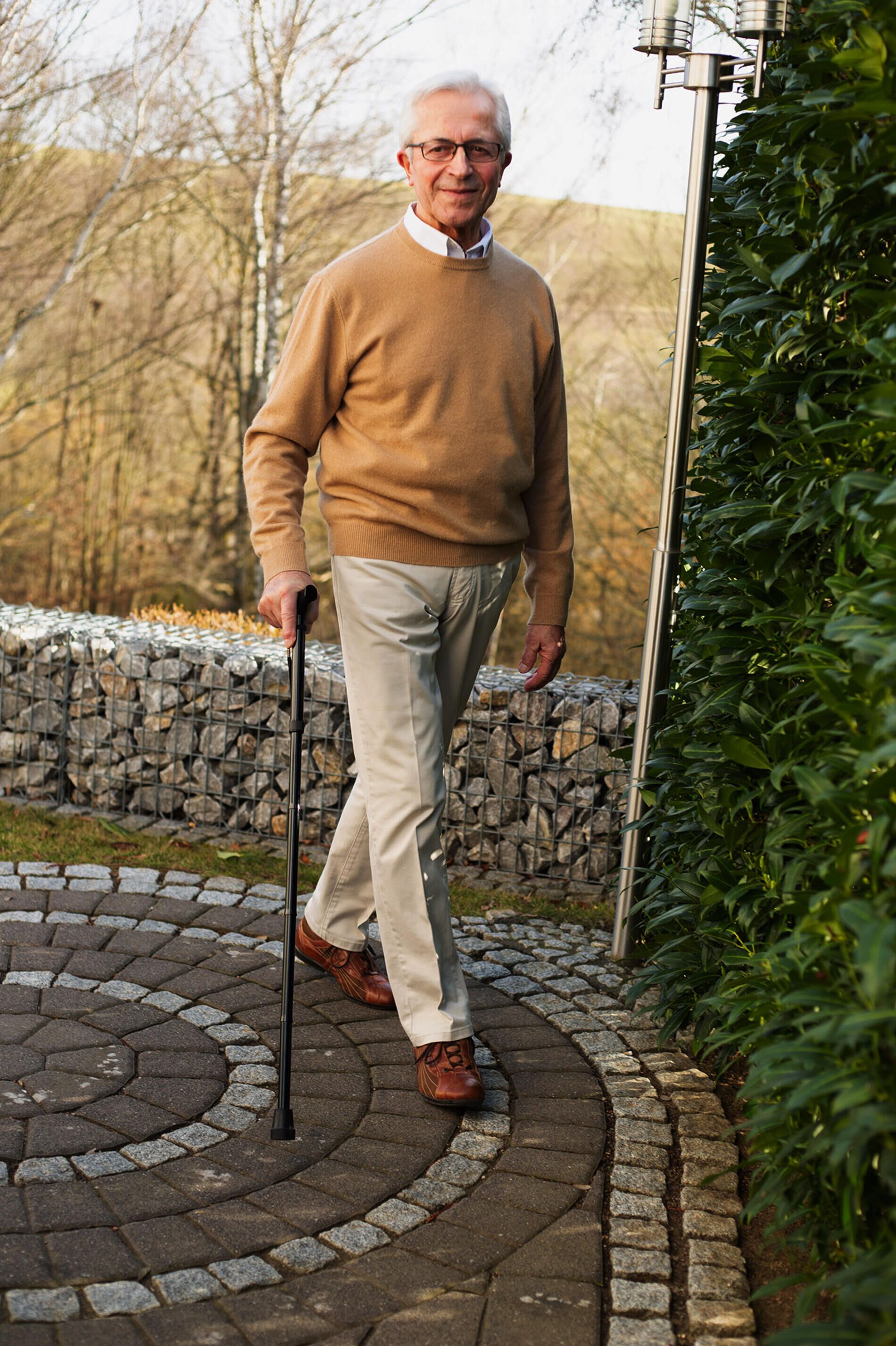 Älterer Mann geht mit schwarzem Gehstock Ally auf einem gepflasterten Weg. Er trägt einen beigen Pullover und eine beige Hose.