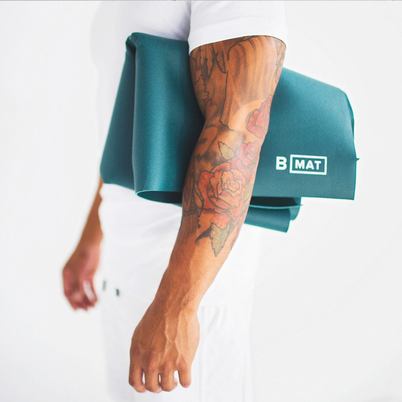 Person trägt gefaltete Yogamatte, dunkelgrün. "B MAT"-Logo sichtbar. Tattoo am Arm.