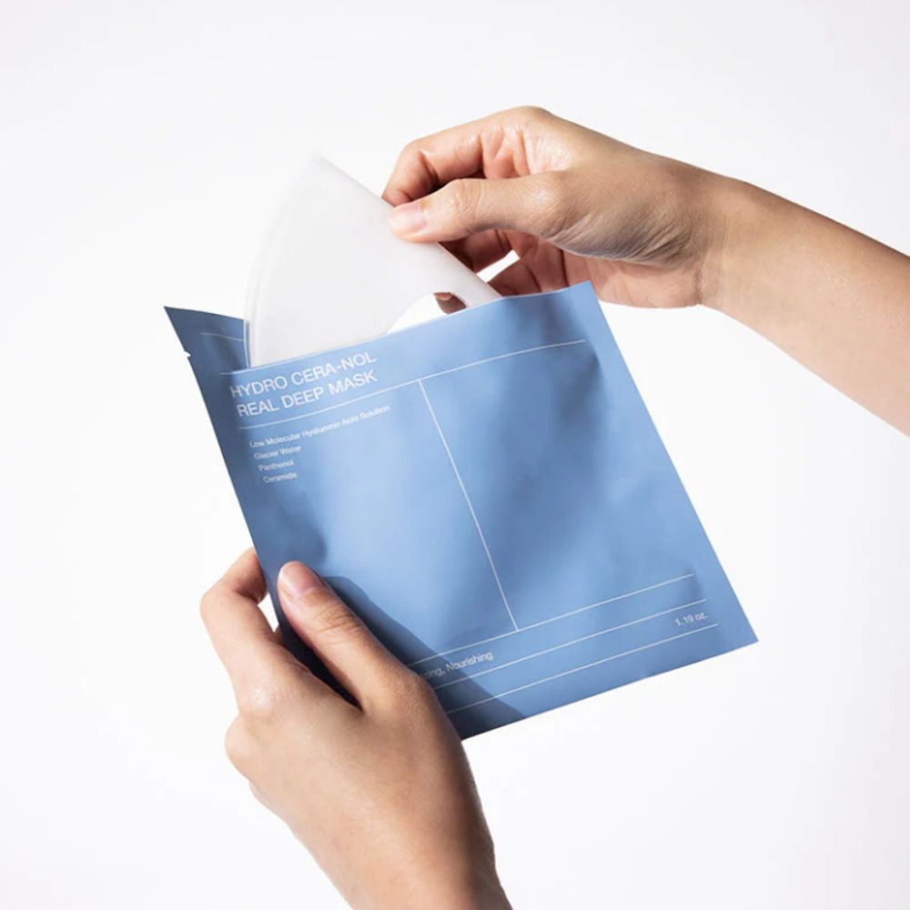 Hände öffnen eine blaue Verpackung. Ein weißes Maskenblatt wird herausgenommen. Auf der Verpackung steht "Biodance Hydro Cera Nol Real Deep Mask".