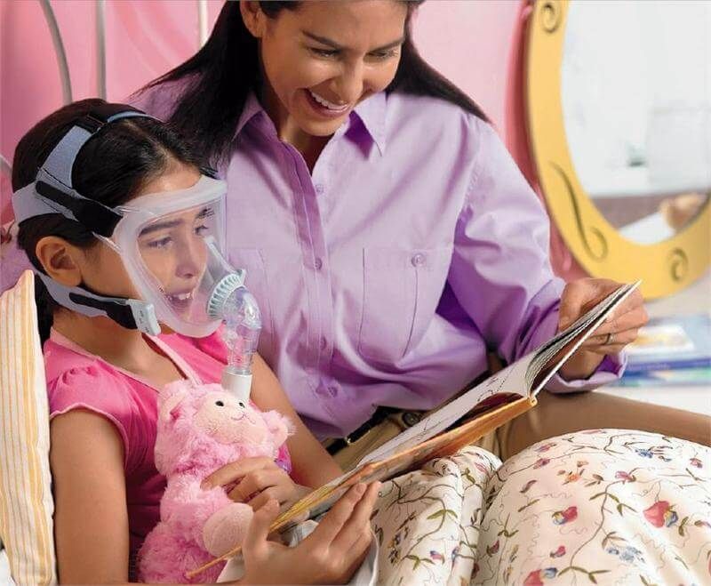 Kind trägt Philips CPAP Vollgesichtsmaske, Mutter liest vor, im Schlafzimmer.