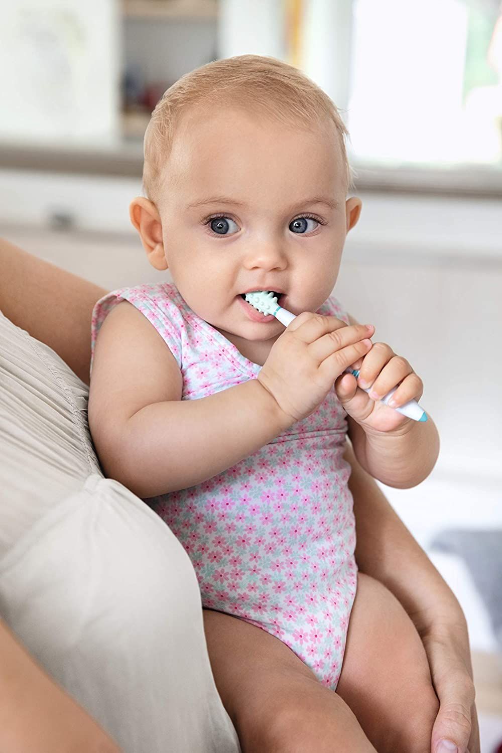 Baby mit Zahnbürste im Mund. Baby trägt einen rosa Strampler. Baby sitzt auf dem Schoß einer Person.