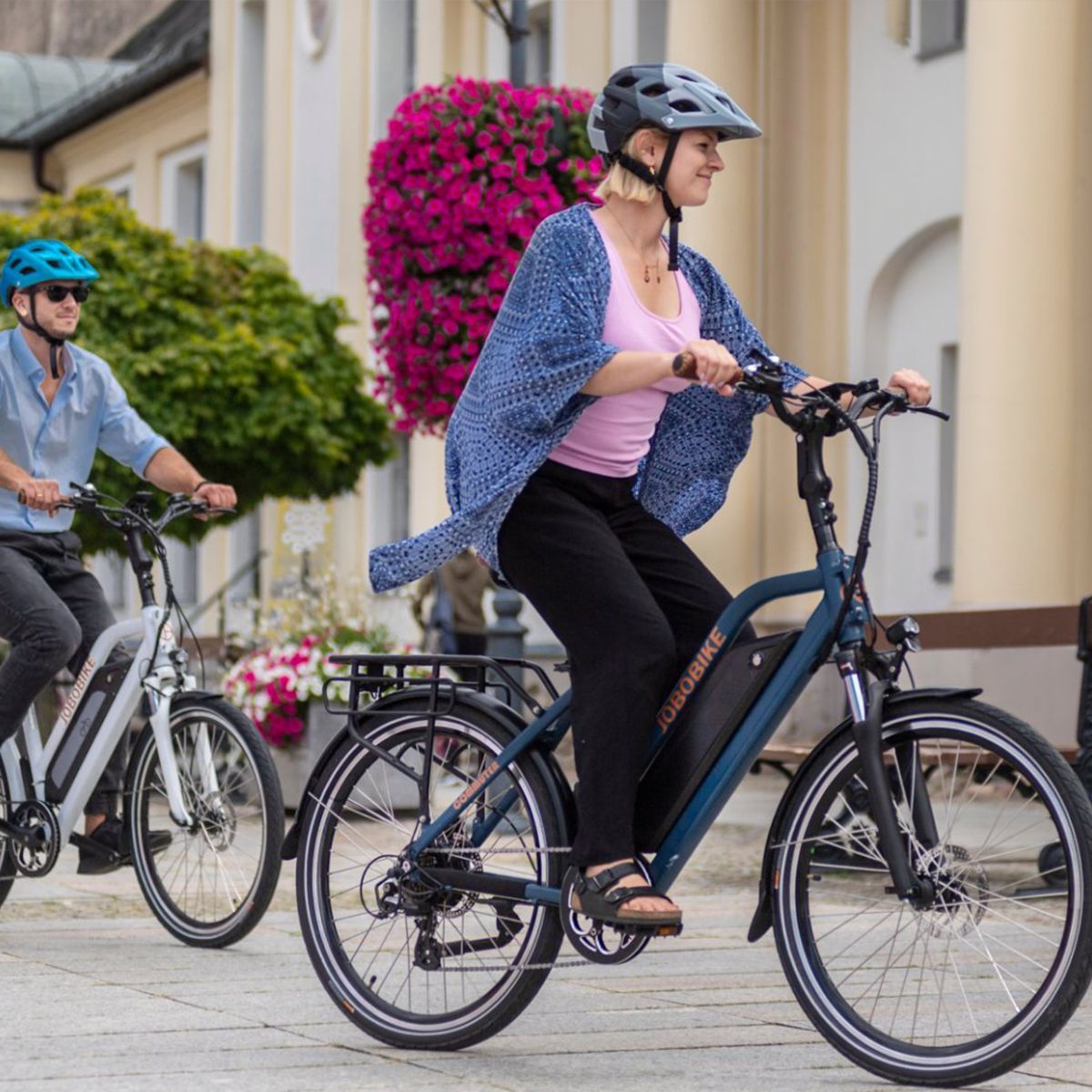 Zwei Personen fahren auf blauen und weißen E-Bikes. Helme, Stadtumgebung.