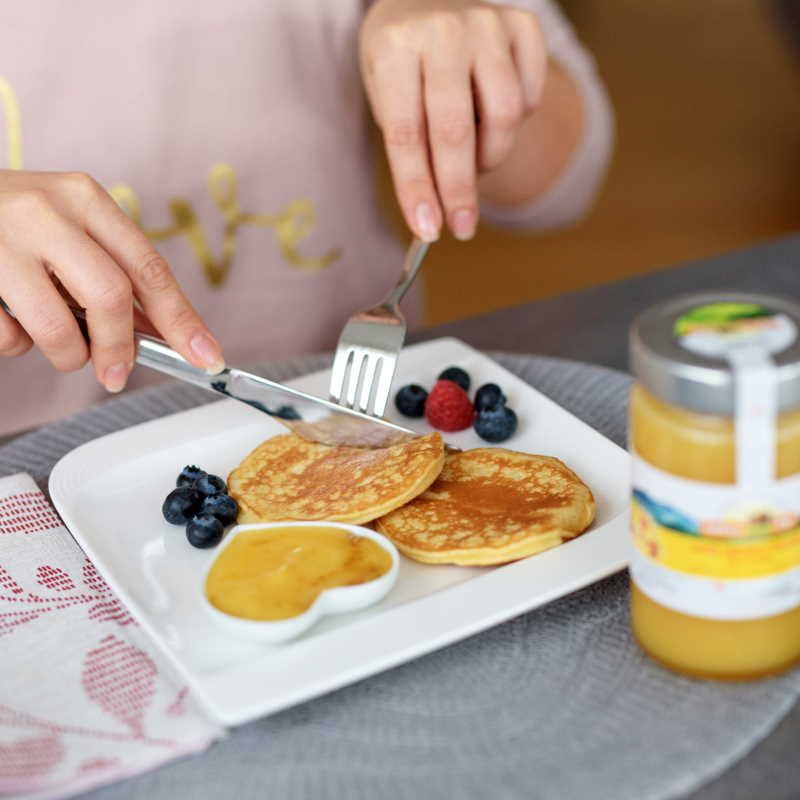 Person schneidet Pfannkuchen mit Honig. Auf dem Teller: Pfannkuchen, Blaubeeren, Honig in Schale. Honigglas rechts.