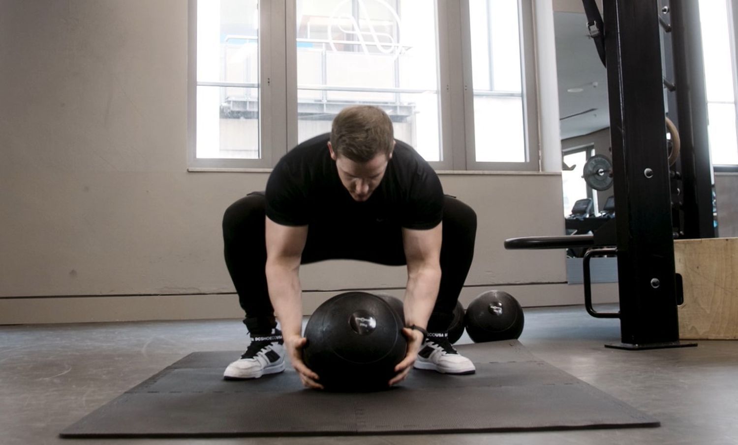 Eine Person in schwarzer Kleidung hockt und hält einen schwarzen Slamball. Im Hintergrund ist ein Fitnessstudio zu sehen.