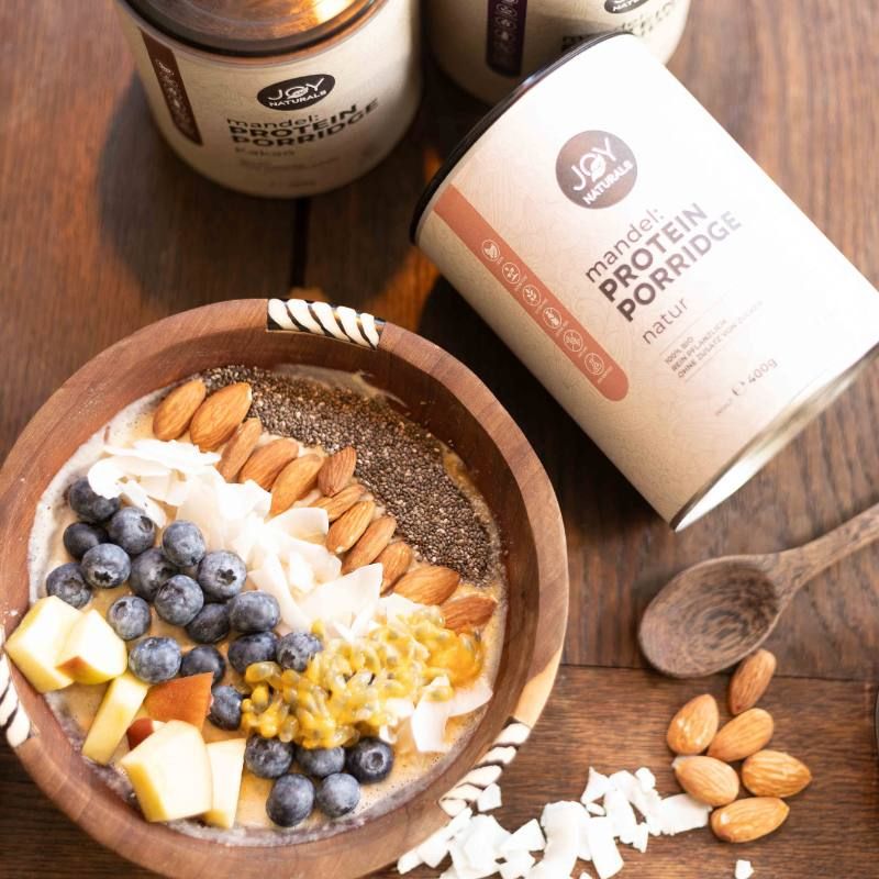 Holzschale mit Porridge, Mandeln, Blaubeeren, Kokosraspeln und Chiasamen. Daneben Dose 'JOY NATURALS'.