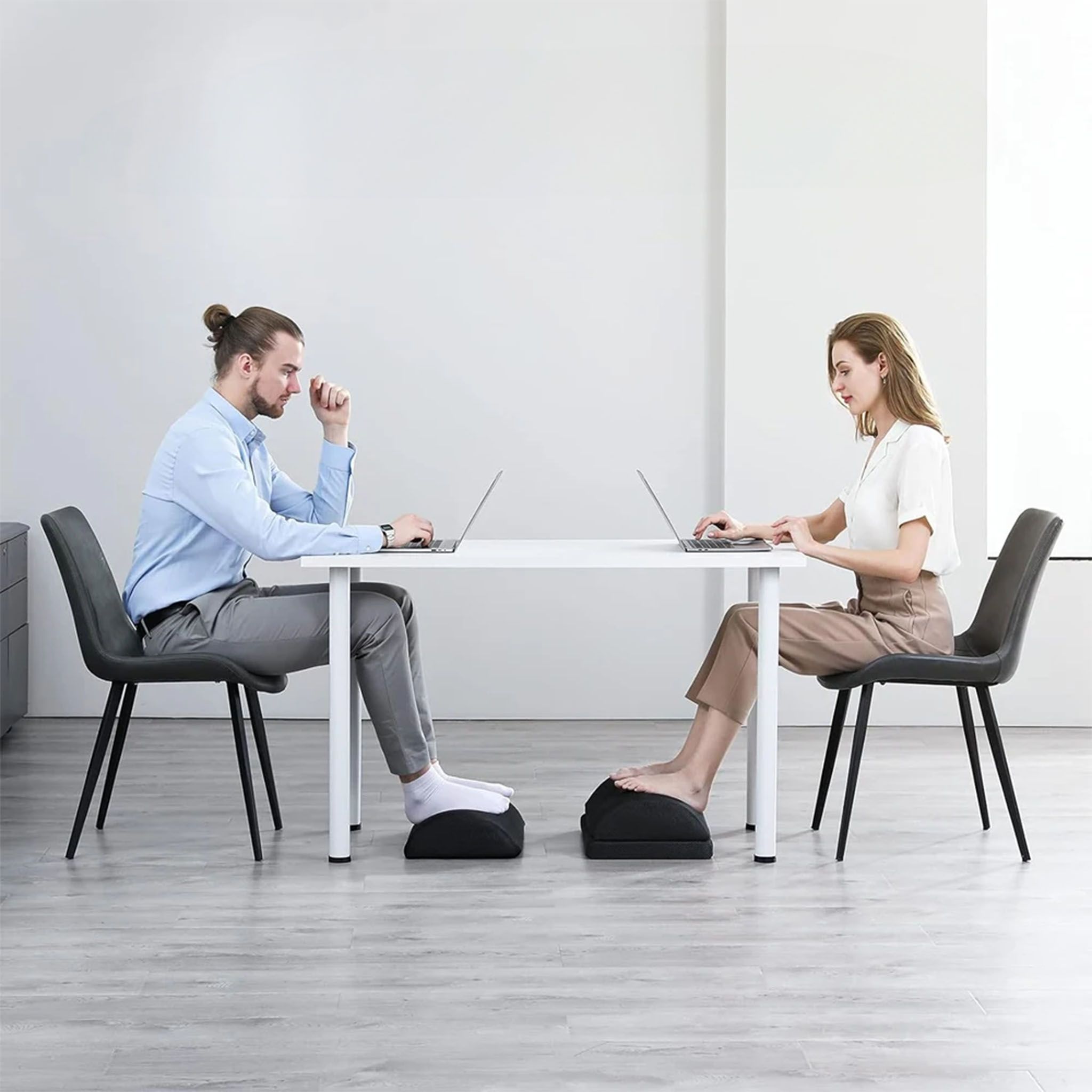 Zwei Personen sitzen an einem Schreibtisch und arbeiten an Laptops. Beide haben einen schwarzen Fußhocker.