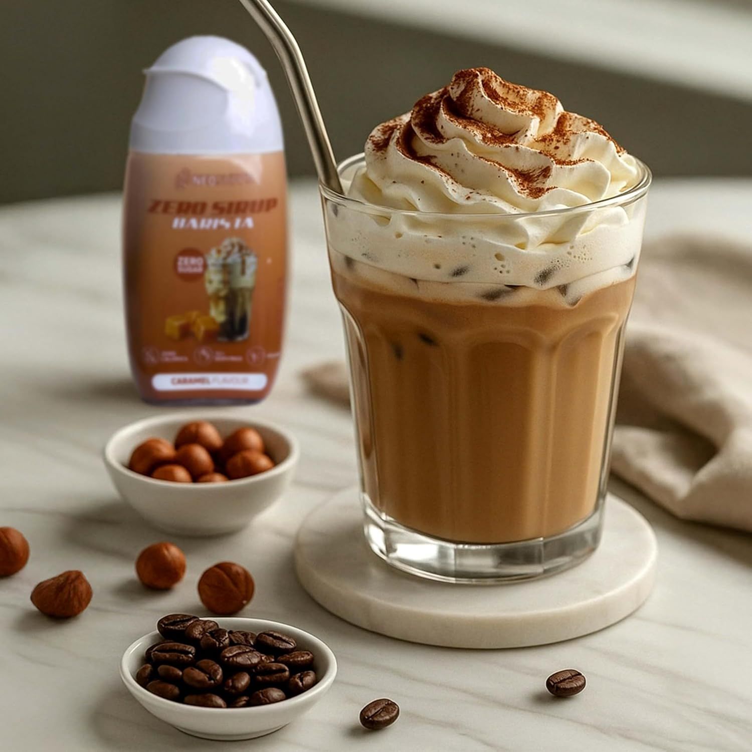 Glas Kaffee mit Sahne, daneben Flasche Neosupps Zero Sirup Barista, Caramel. Haselnüsse und Kaffeebohnen in Schalen. Edelstahl-Trinkhalm.