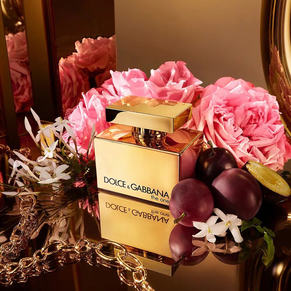 Goldfarbener Flakon mit Aufschrift Dolce & Gabbana The One. Umgeben von Blumen, Früchten und Schmuck.