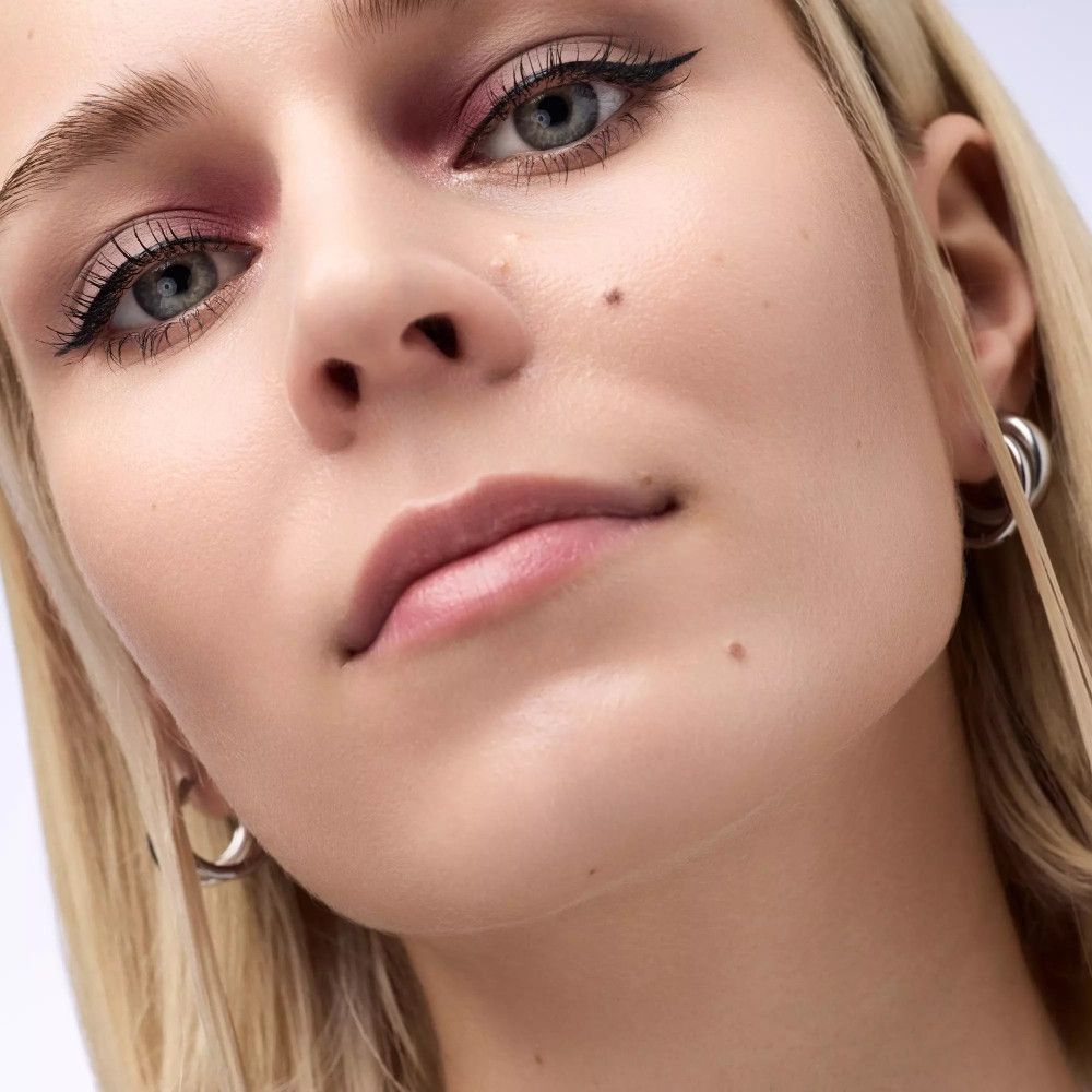 Porträt einer Person mit blonden Haaren und rosa Lidschatten. Schwarzer Eyeliner, silberne Ohrringe.