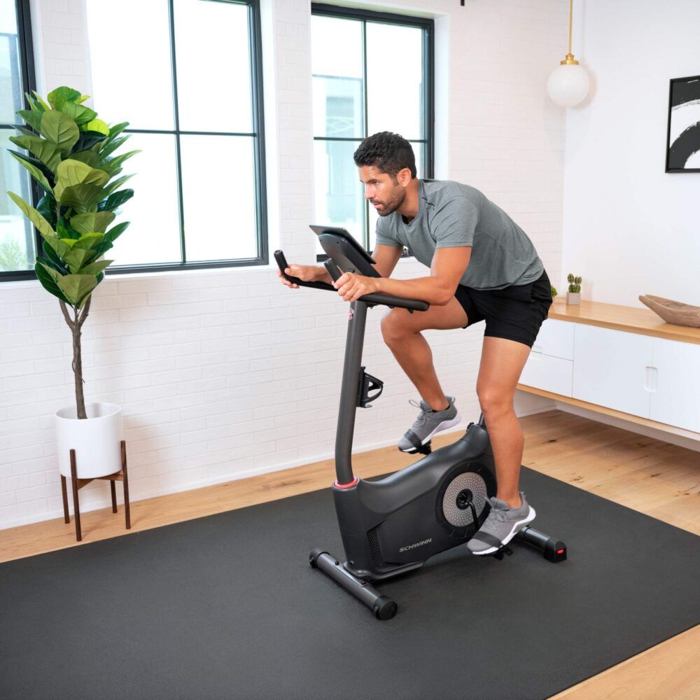 Mann auf einem schwarzen Hometrainer. Schwinn-Logo. In einem Wohnzimmer. Schwarze Matte. Graue Kleidung.