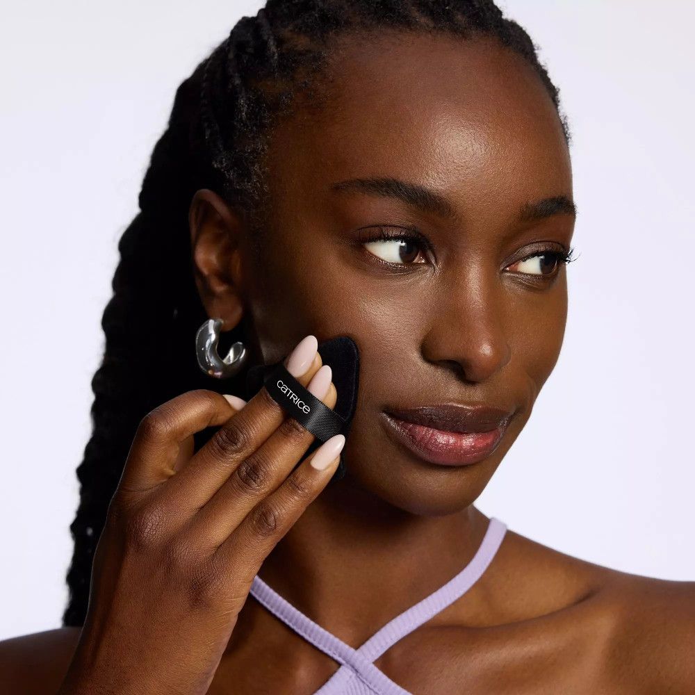 Schwarzes Catrice Powder Puff wird auf der Wange verwendet. Dreieckige Form mit schwarzem Band und Markenname.