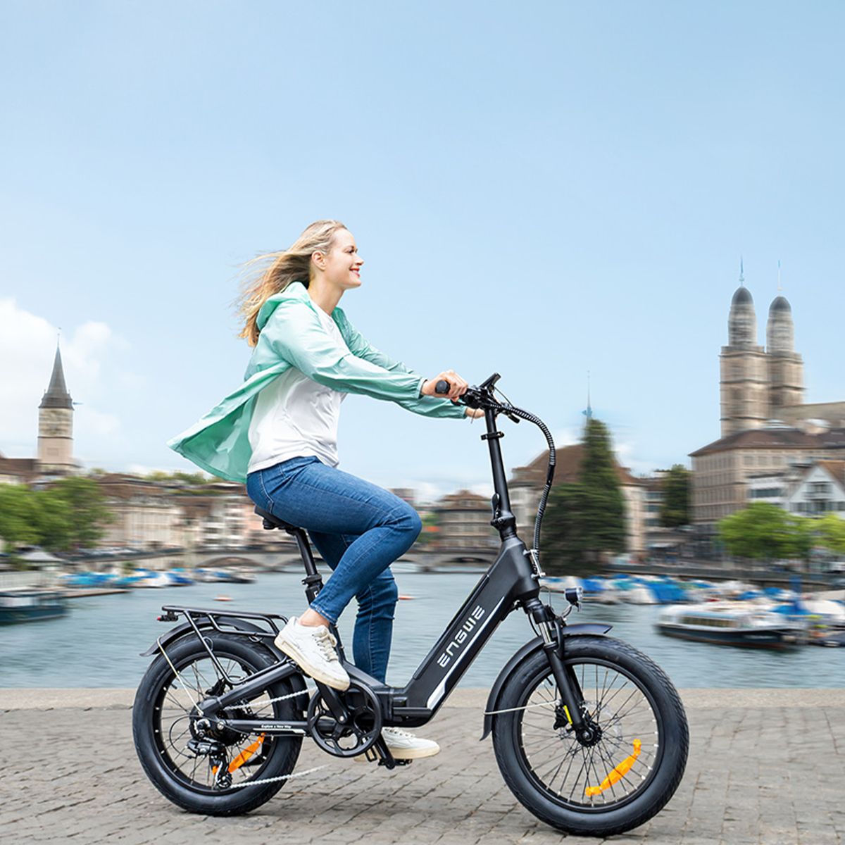 Frau fährt auf einem schwarzen Elektrofahrrad. Das ENGWE-Logo ist sichtbar. Hintergrund: Gebäude und Wasser.