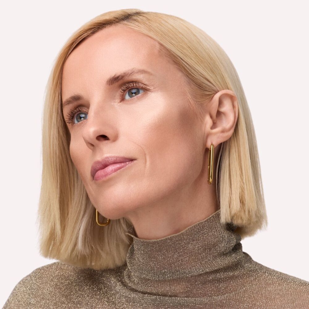 Porträt einer Frau mit blonden Haaren und goldenem Rollkragen. Highlighter auf Wangenknochen und Stirn. Goldene Ohrringe.