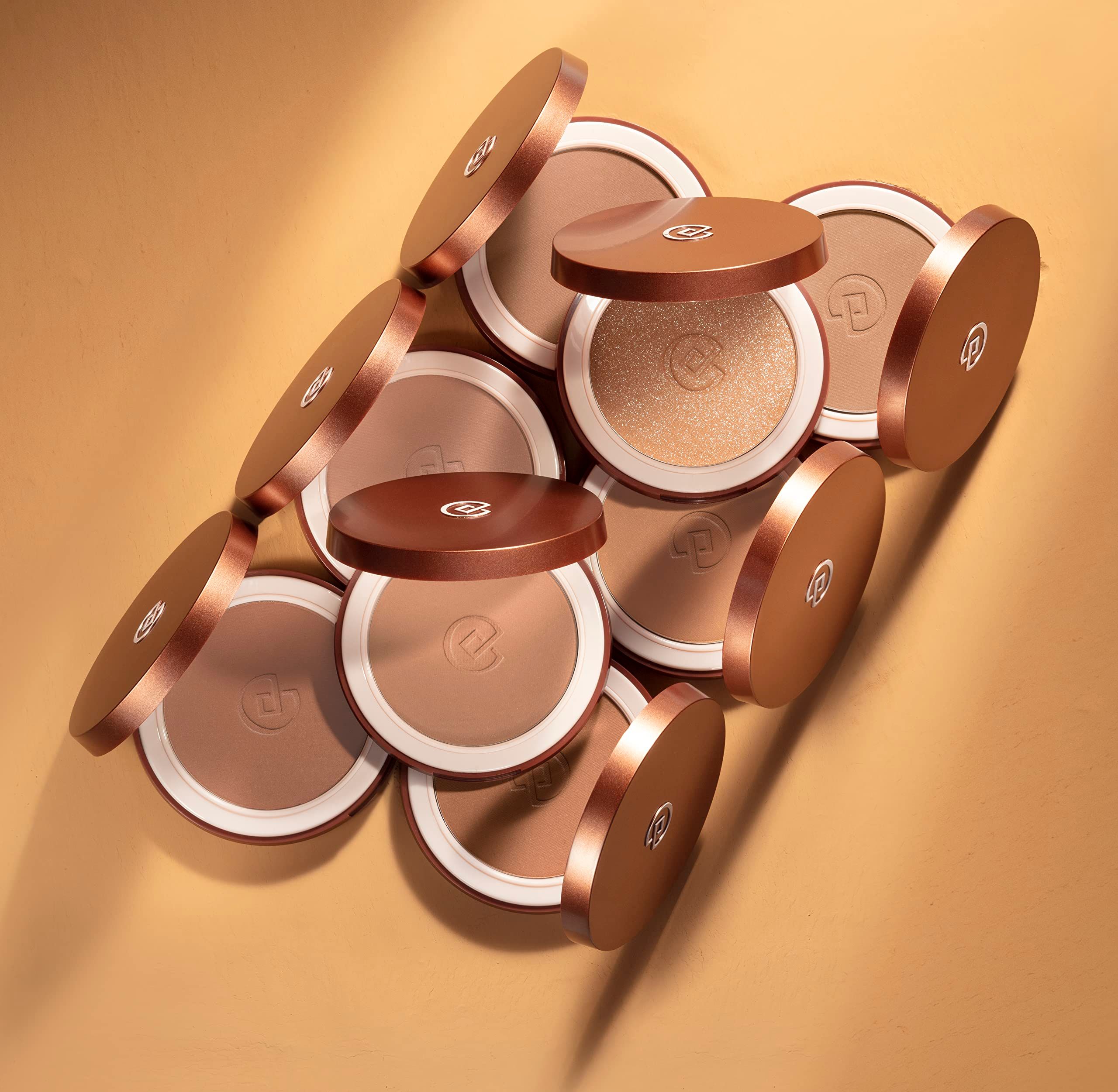 Mehrere Bronzer mit geöffneten Deckeln. Unterschiedliche Farbtöne. Geprägtes Logo. Auf gelbem Hintergrund.