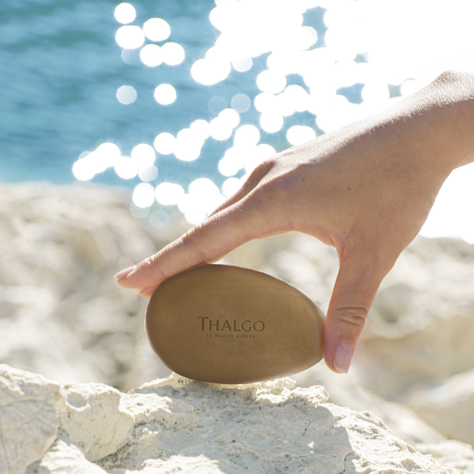 Hand hält ovale Seife mit THALGO-Logo. Seife auf weißem Felsen, Hintergrund Meer und Sonne.