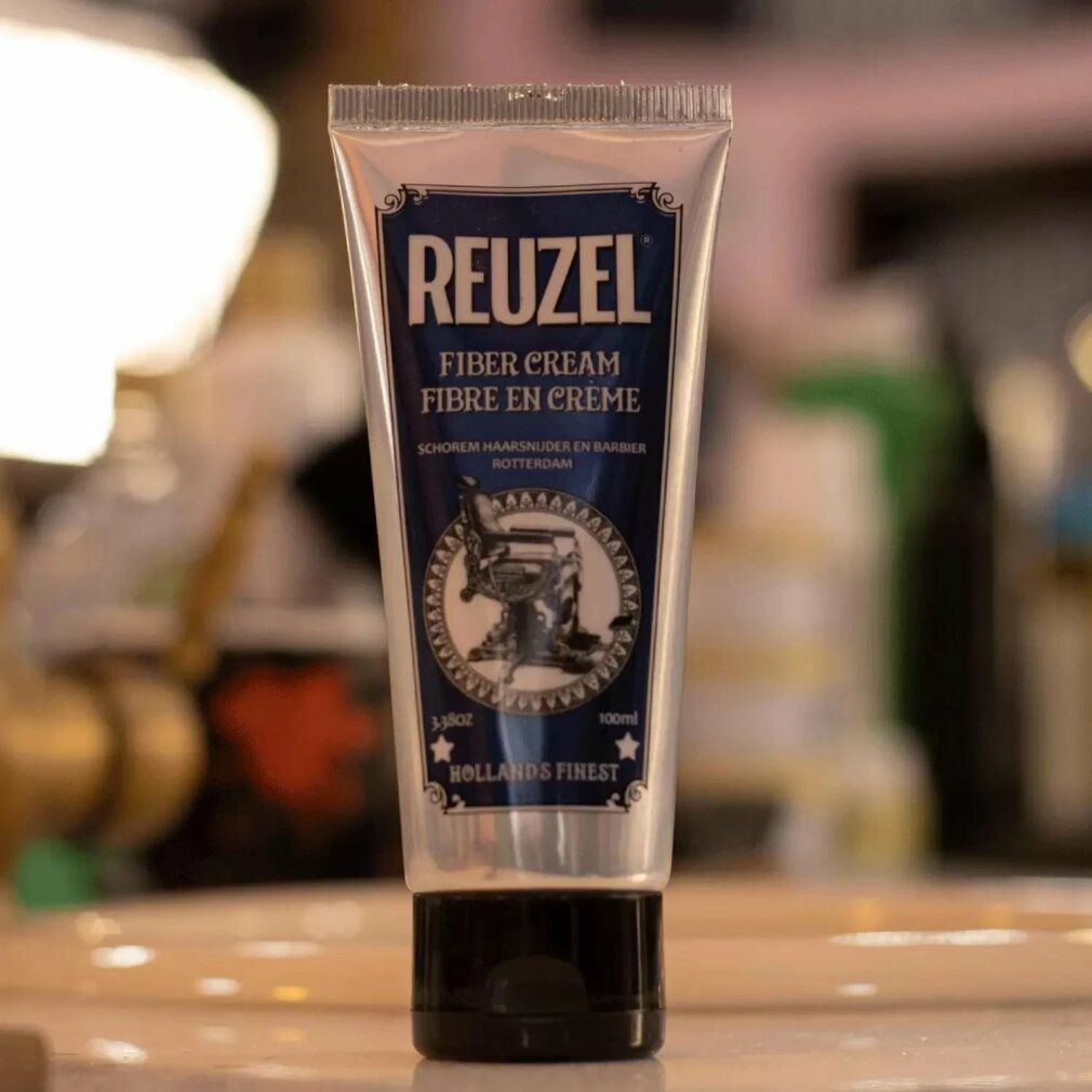 Silberne Tube mit blauem Etikett. Aufschrift REUZEL FIBER CREAM. Abbildung eines Friseurstuhls. Schwarzer Schraubverschluss.