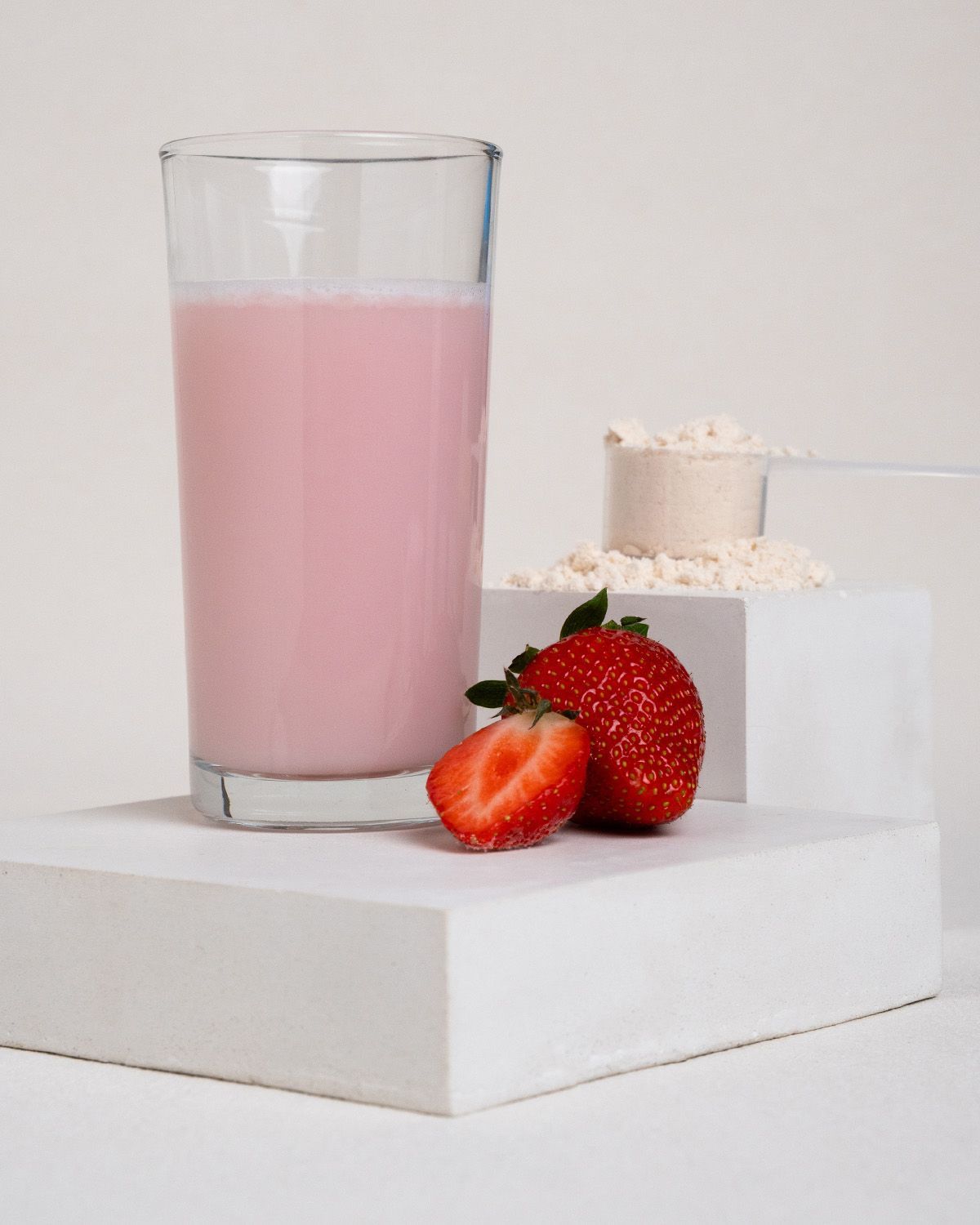 Glas rosa Flüssigkeit, daneben Erdbeeren und Proteinpulver mit Messlöffel. Auf weißen Blöcken angeordnet.