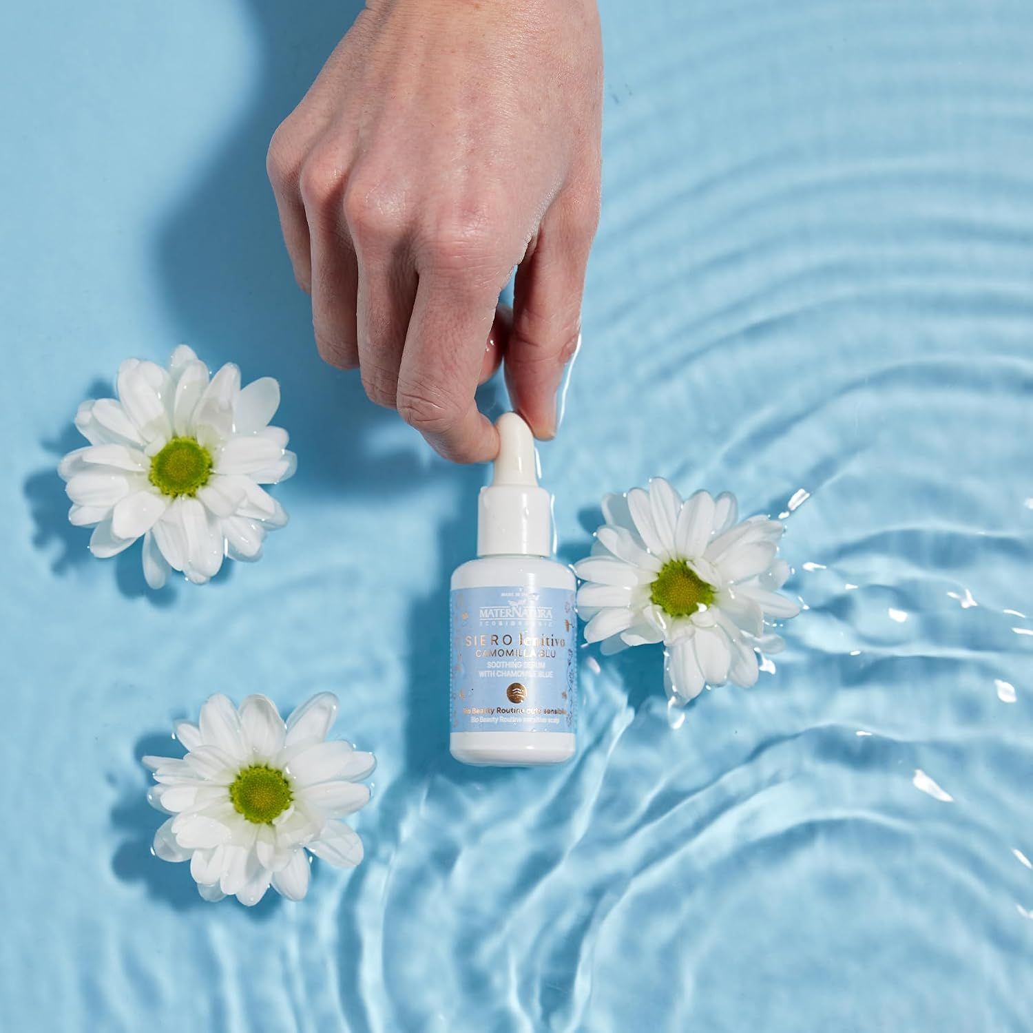 Eine Hand hält einen kleinen Flakon mit Pipette über Wasser mit Blüten.