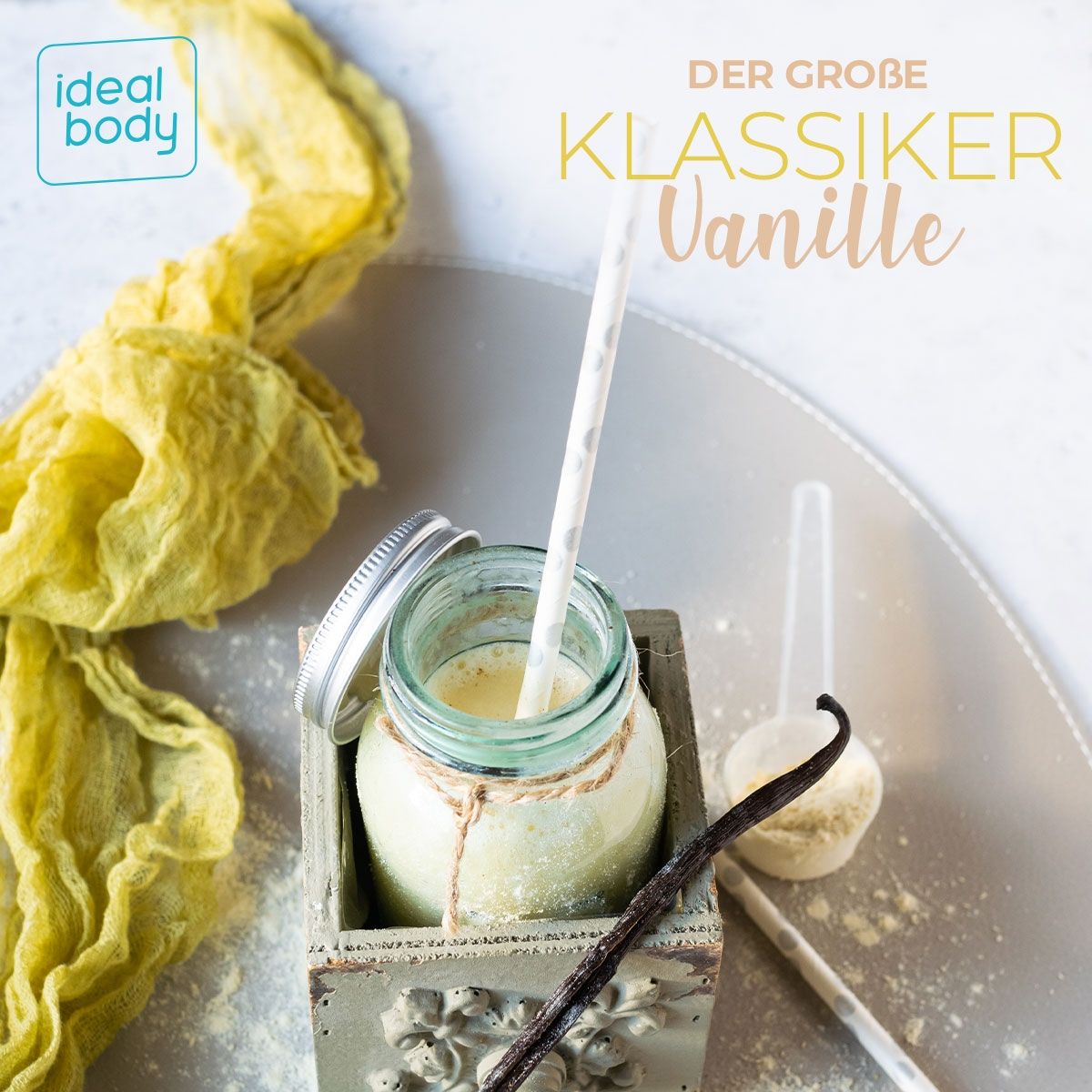 Glas mit Vanille-Proteinshake, Strohhalm, Vanilleschote und Löffel. Aufschrift: "DER GROßE KLASSIKER Vanille". Gelbes Tuch im Hintergrund.
