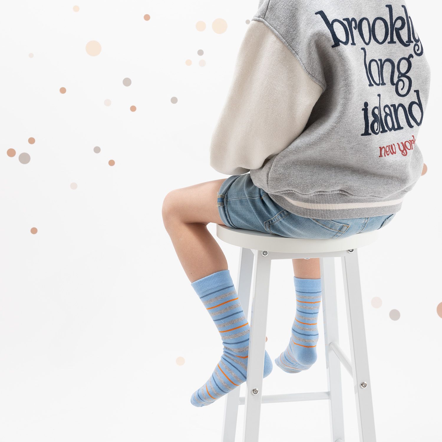 Kind sitzt auf einem Hocker, trägt gestreifte Socken in Blau und Orange. Kurze Hose und Jacke.