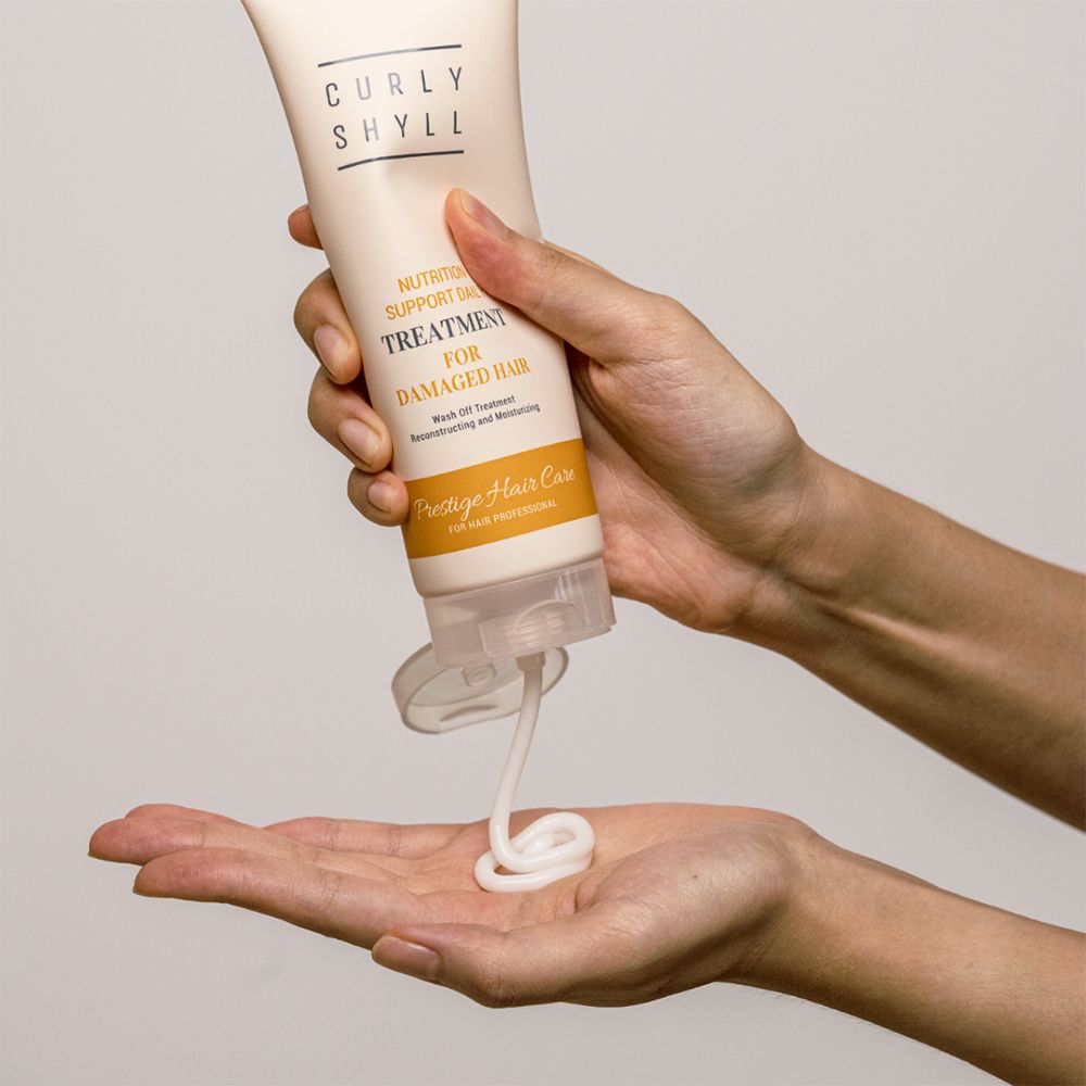Hände halten eine Tube. Creme wird herausgedrückt. Aufschrift: CURLY SHYLL, TREATMENT FOR DAMAGED HAIR. Untertitel: Prestige Hair Care.