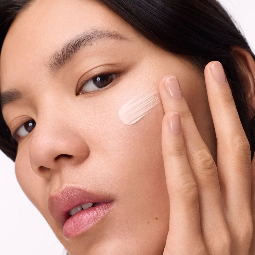 Frau trägt Primer auf. Gesicht einer Frau, die Primer auf die Wange aufträgt. Hautton-Primer wird mit dem Finger aufgetragen.