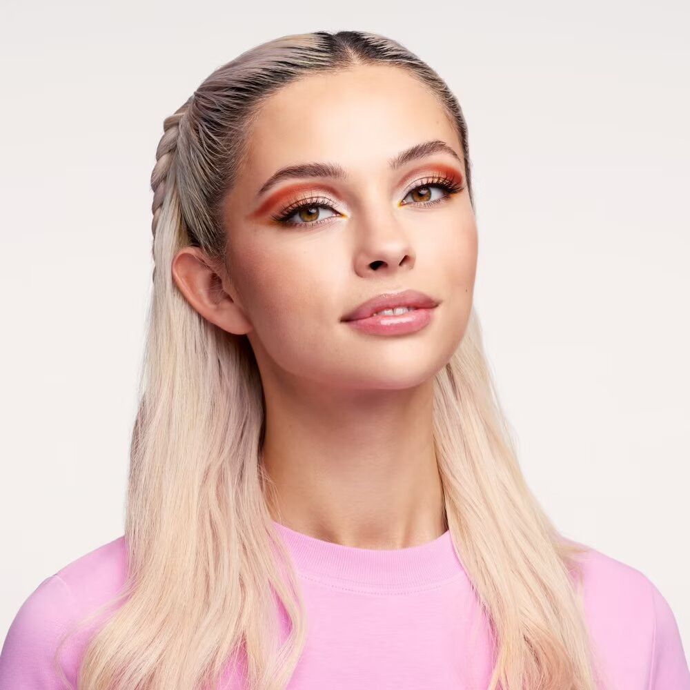 Porträt einer Frau mit blonden Zöpfen. Sie trägt ein pinkfarbenes Oberteil. Ihr Make-up umfasst Lidschatten und Lipgloss.