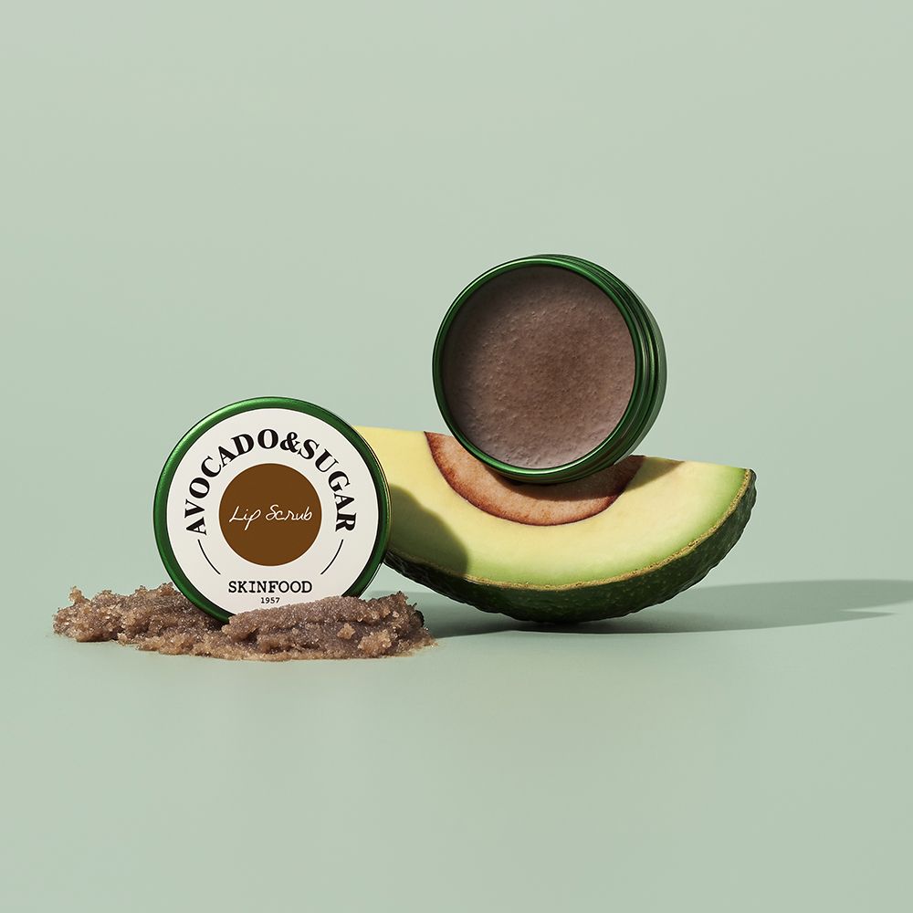 Zwei grüne Behälter, einer geöffnet, neben einer Avocado-Hälfte und einem Haufen Scrub. Aufschrift: Avocado & Sugar, SKINFOOD.