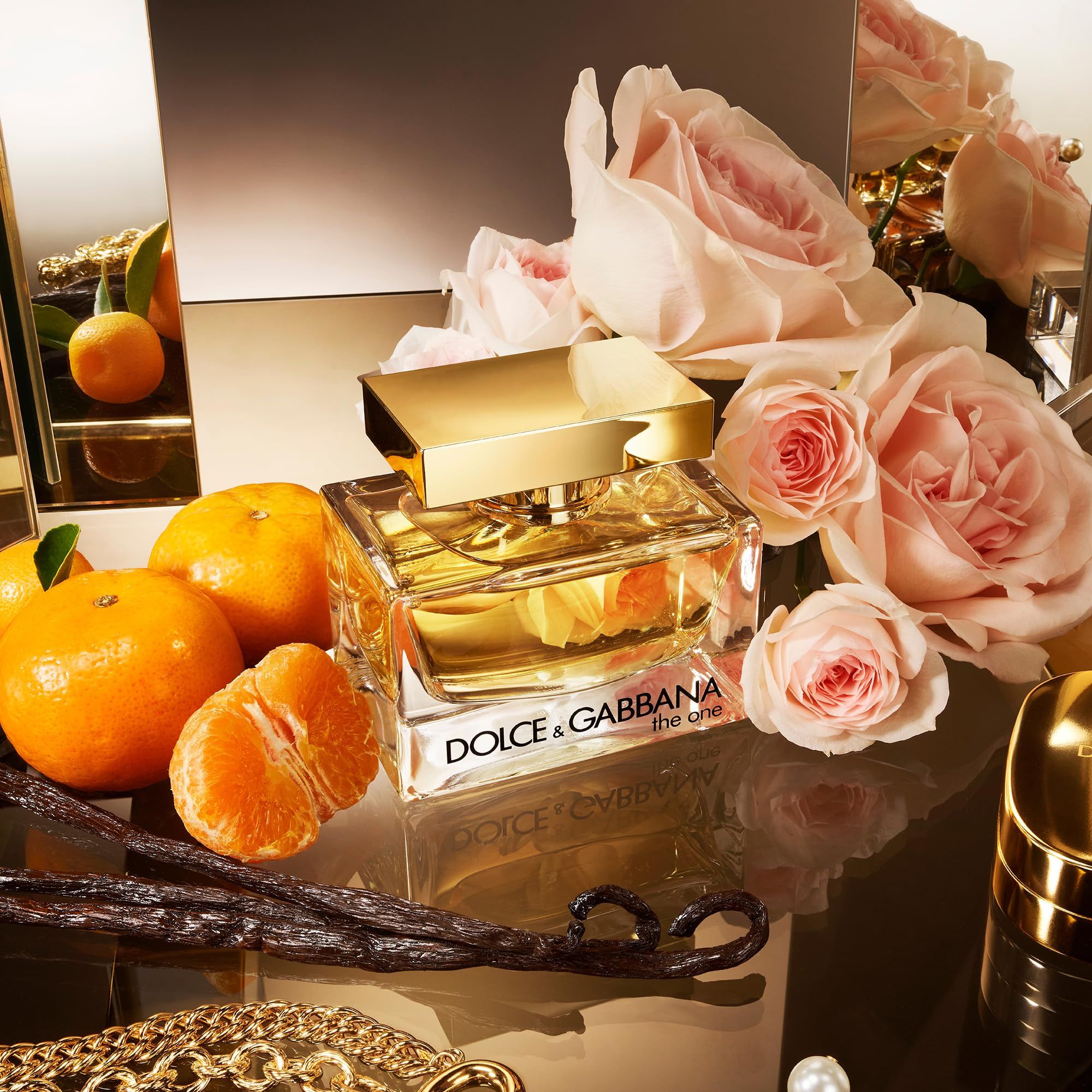 Parfümflakon, goldfarbener Verschluss, umgeben von Rosen, Mandarinen und Vanilleschoten. Spiegelung auf glänzender Oberfläche.
