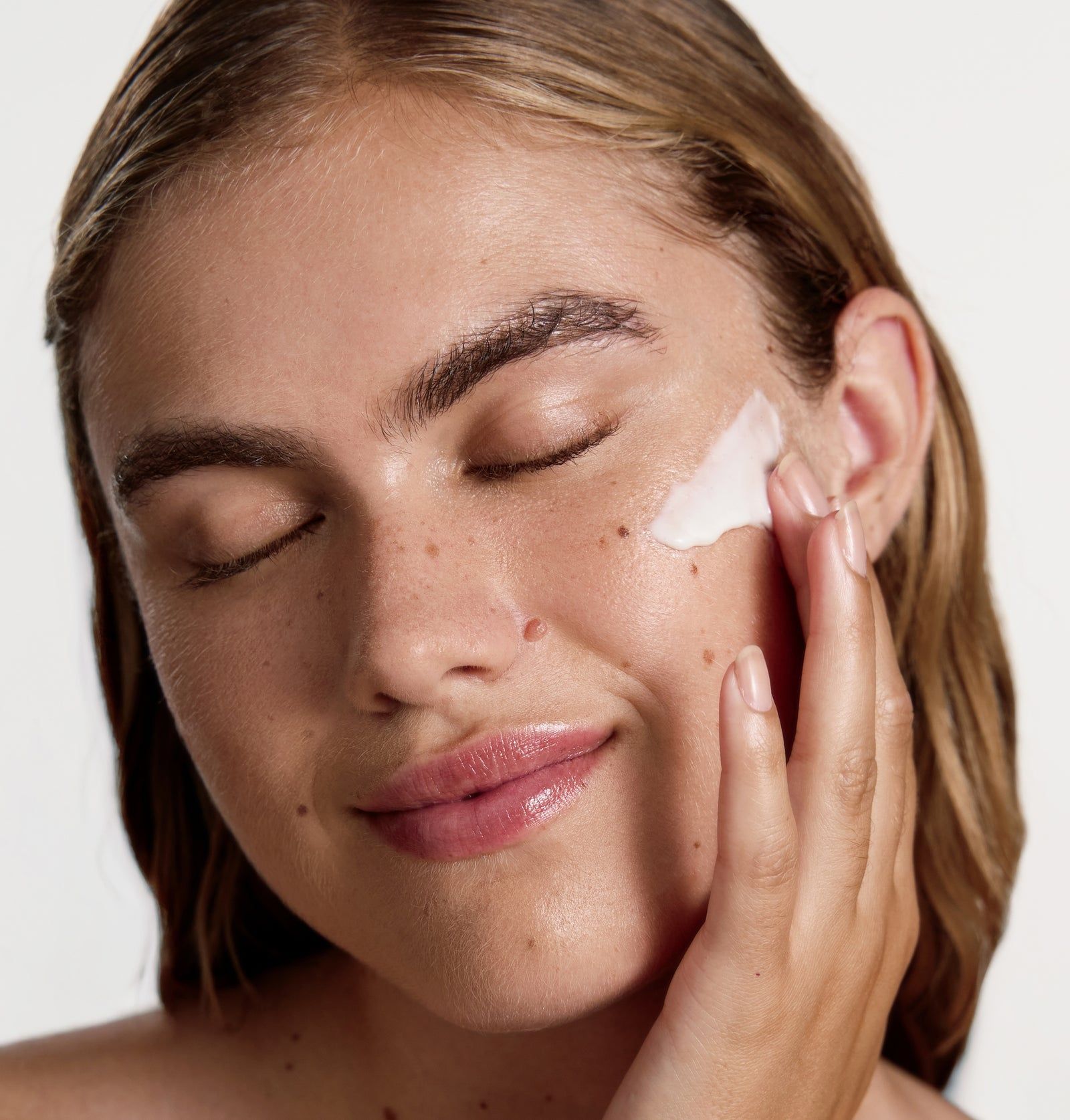 Frau mit geschlossenen Augen, Creme auf der Wange. Helle Haut, braune Haare.