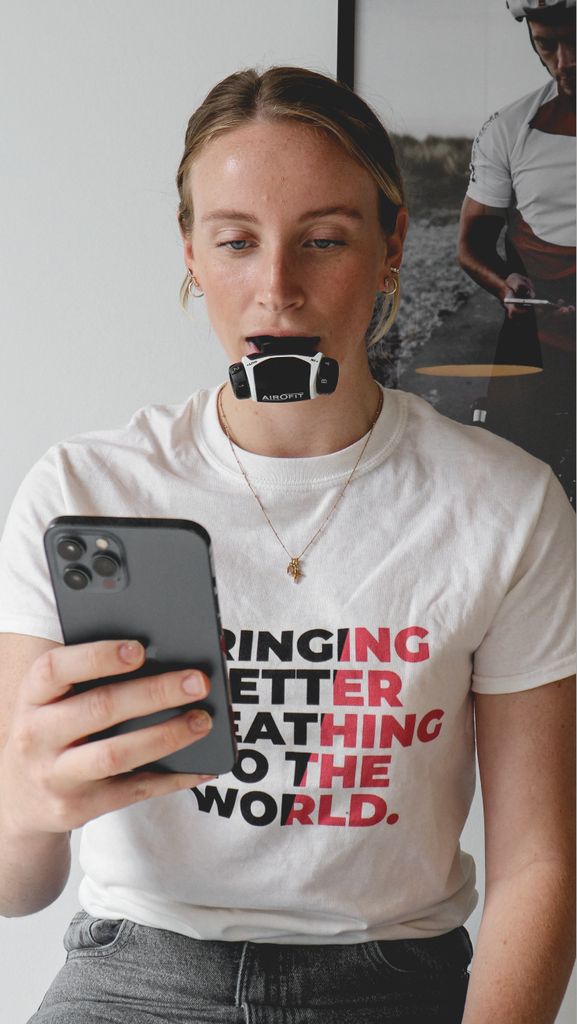 Frau mit Atemtrainer im Mund, hält Smartphone. 'BRINGING BETTER BREATHING TO THE WORLD' auf T-Shirt.