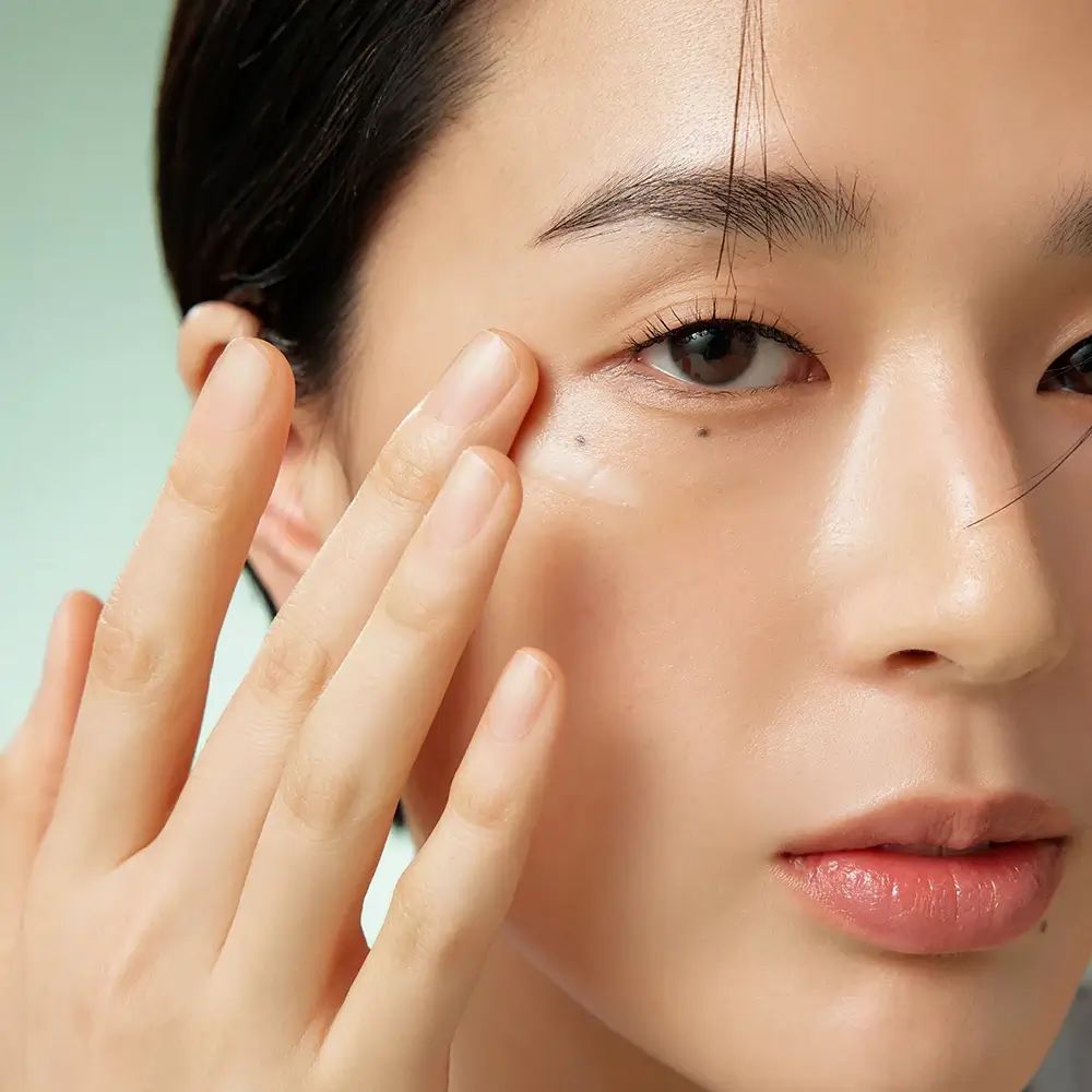 Nahaufnahme einer Person, die Creme unter dem Auge aufträgt. Die Hand berührt sanft die Haut. Helle Haut, dunkle Haare.