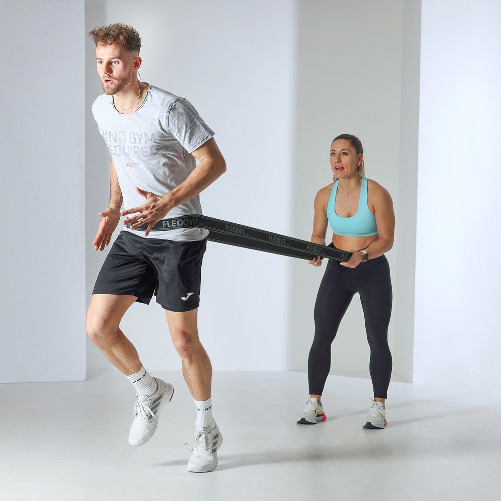 Mann trainiert mit FLEXVIT Resist Band. Frau hält das Band. Graues T-Shirt, schwarze Shorts, blaue Sport-BH. Band schwarz.