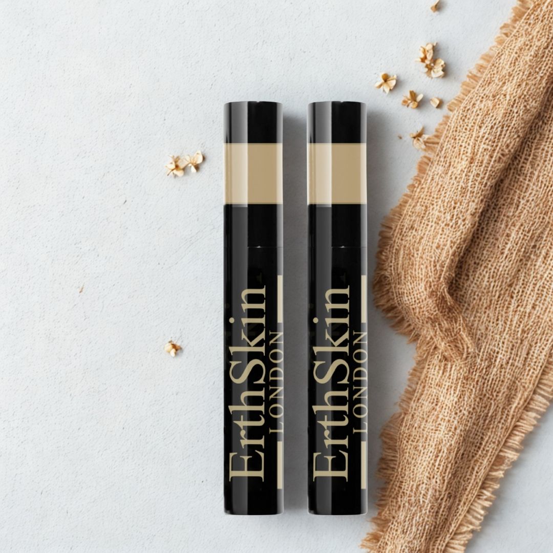 Zwei schwarze Mascara-Tuben mit goldfarbenem Text. Aufschrift: ErthSkin London. Beigefarbener Bereich am oberen Ende. Auf hellem Untergrund mit Stoff und Blüten.