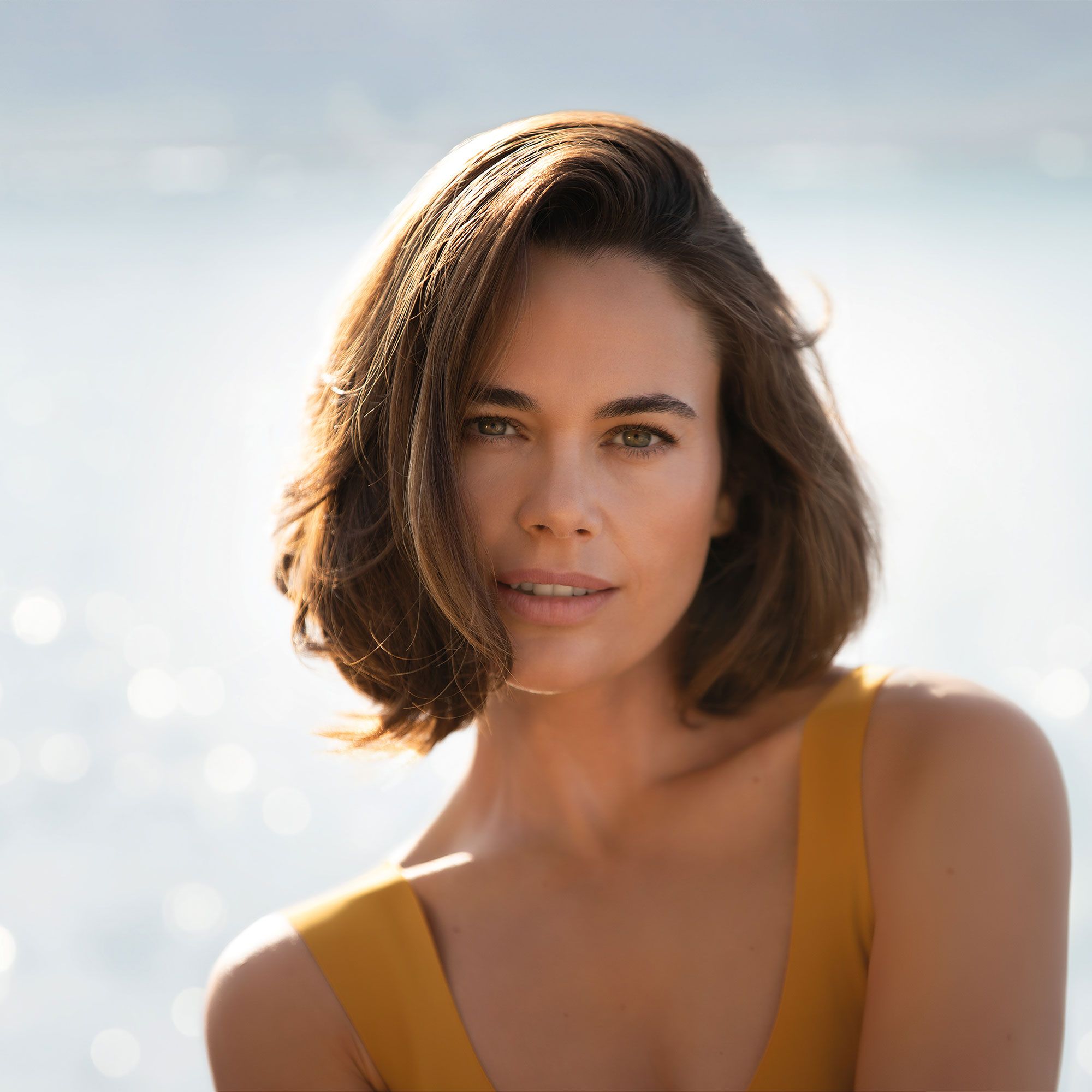Porträt einer Frau. Kurze, braune Haare. Gelbes Oberteil. Blick zur Kamera. Hintergrund: Meer.