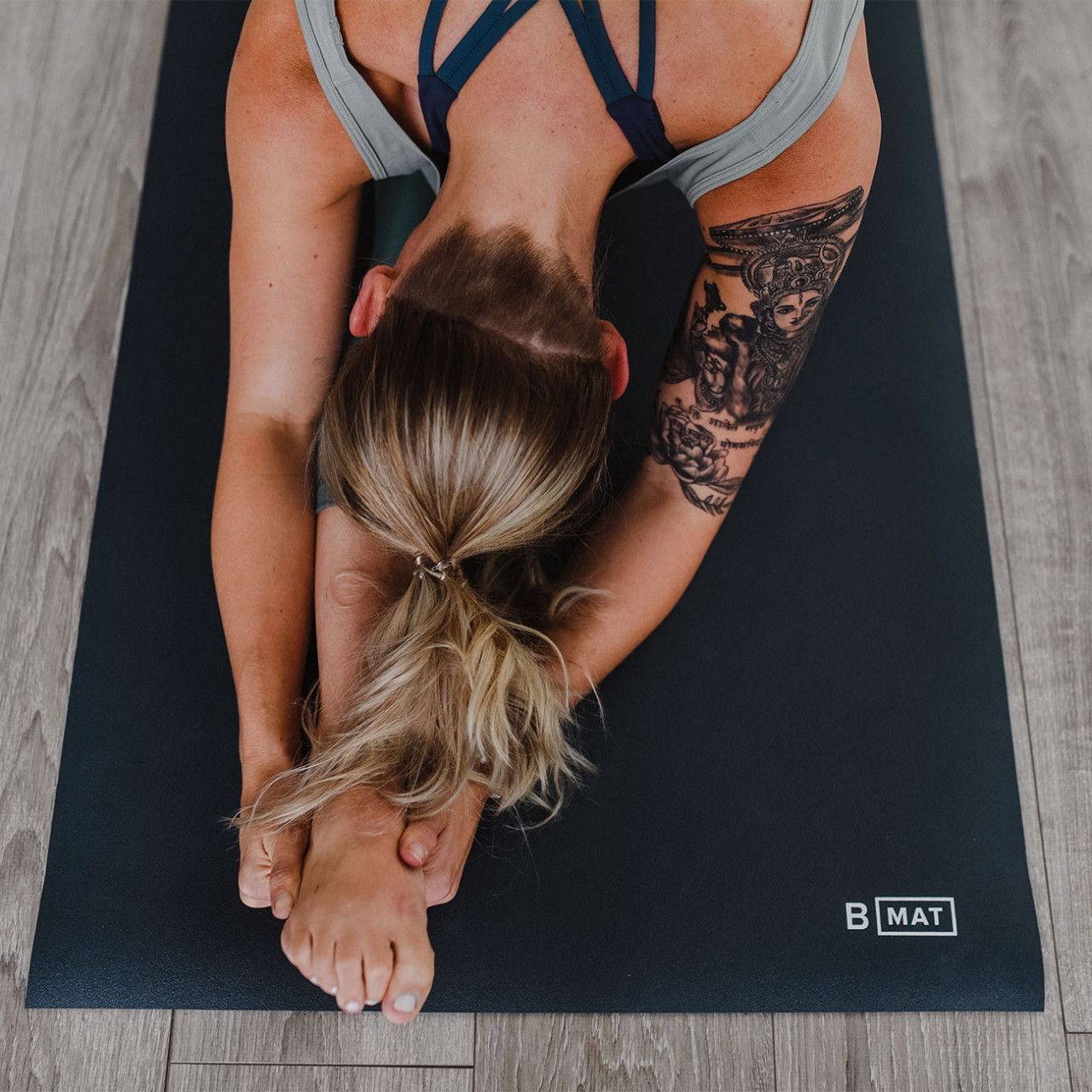 Person praktiziert Yoga auf grauer Yogamatte. Tattoo auf Arm sichtbar. Logo "B MAT".