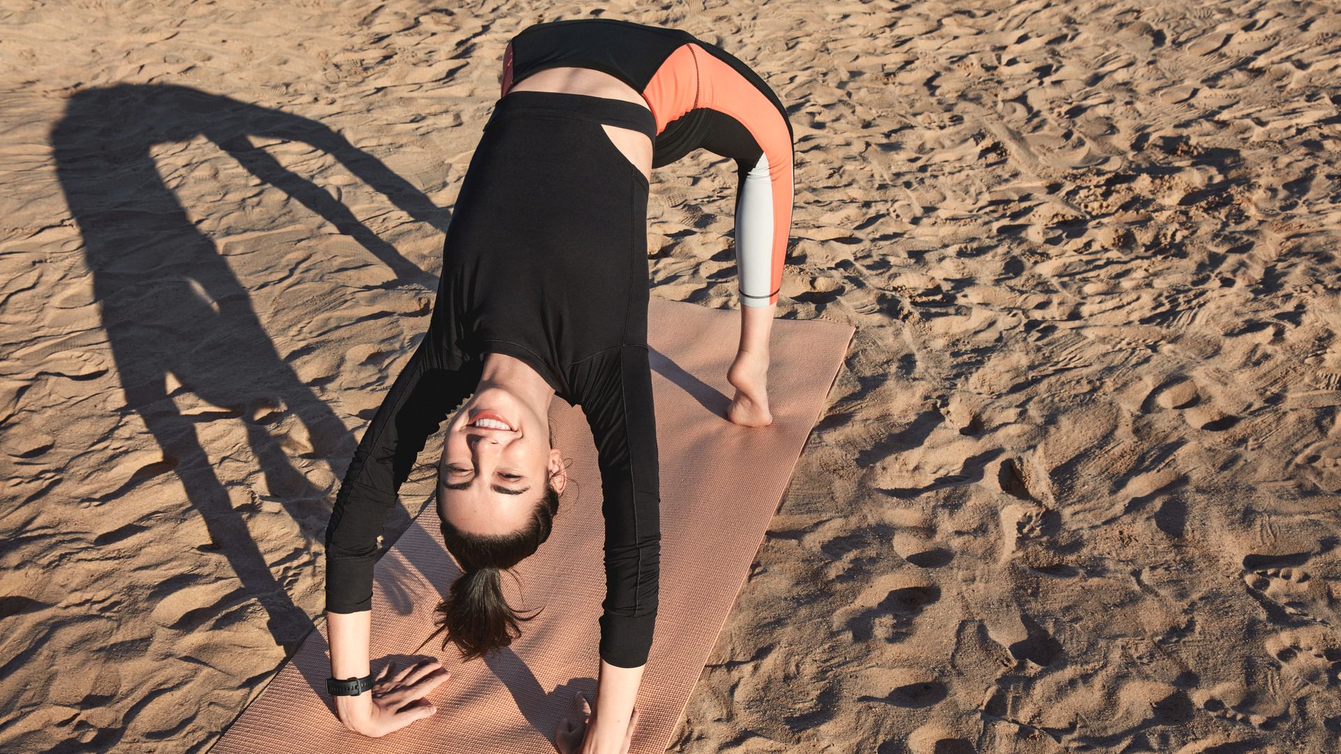 Eine Person macht Yoga auf einer Matte am Strand. Sie trägt Sportkleidung und eine Smartwatch am Handgelenk.