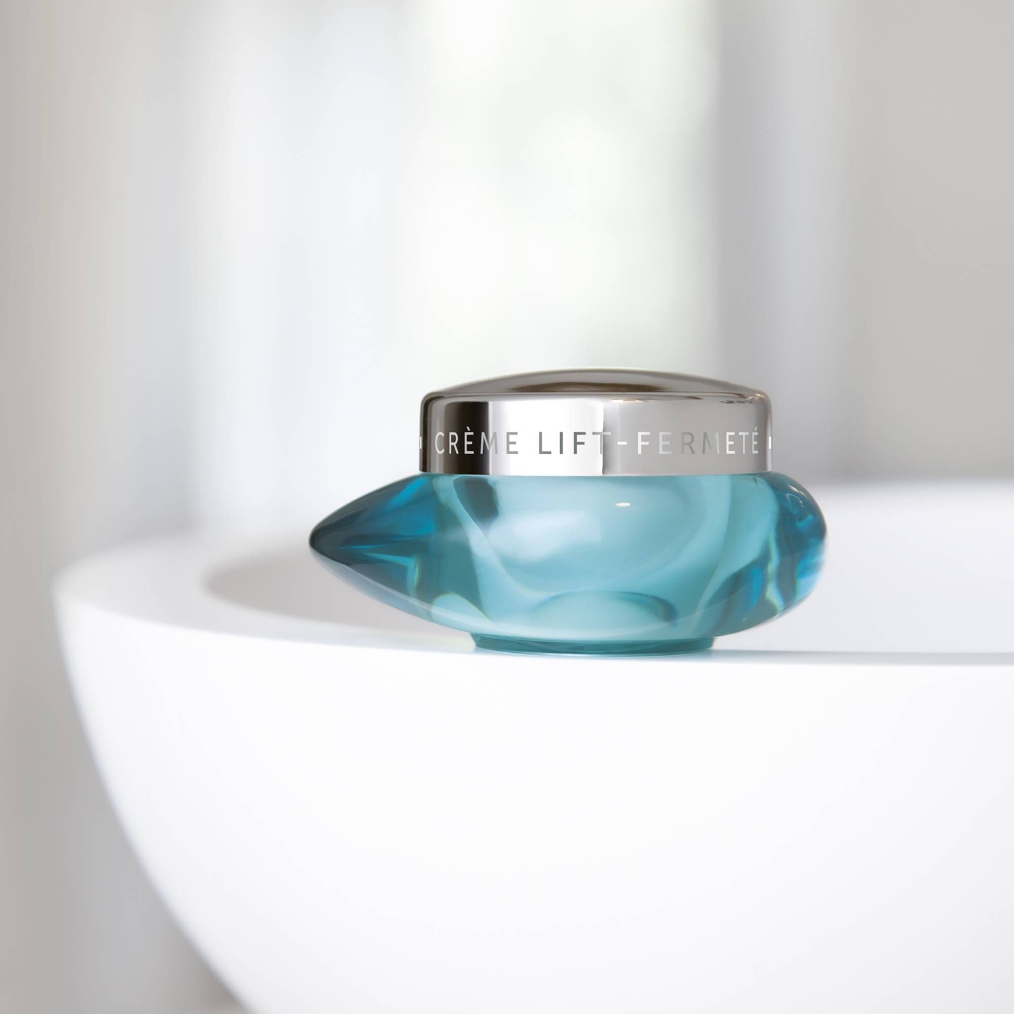 Türkisfarbener Tiegel mit silbernem Deckel. Aufschrift: CRÈME LIFT-FERMETÉ. Auf weißer Oberfläche platziert.