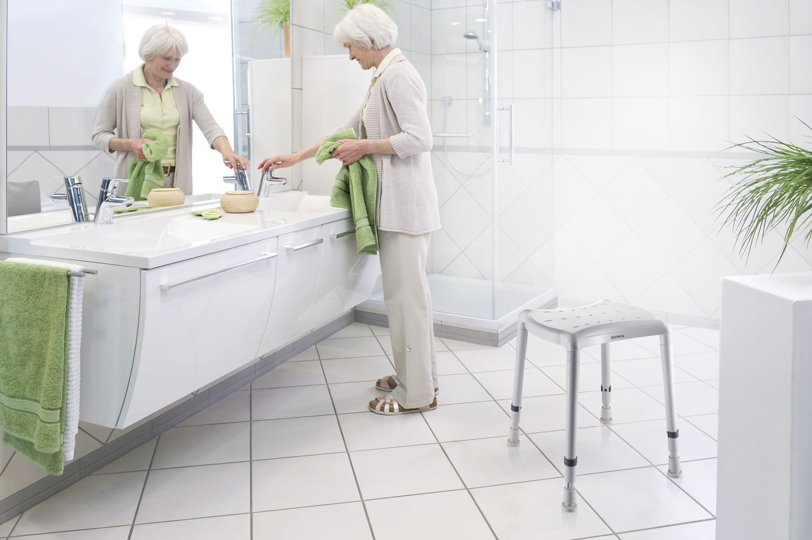 Ältere Frau in einem Badezimmer. Sie steht vor einem Waschbecken und neben einem Duschhocker. Das Badezimmer ist hell und modern.