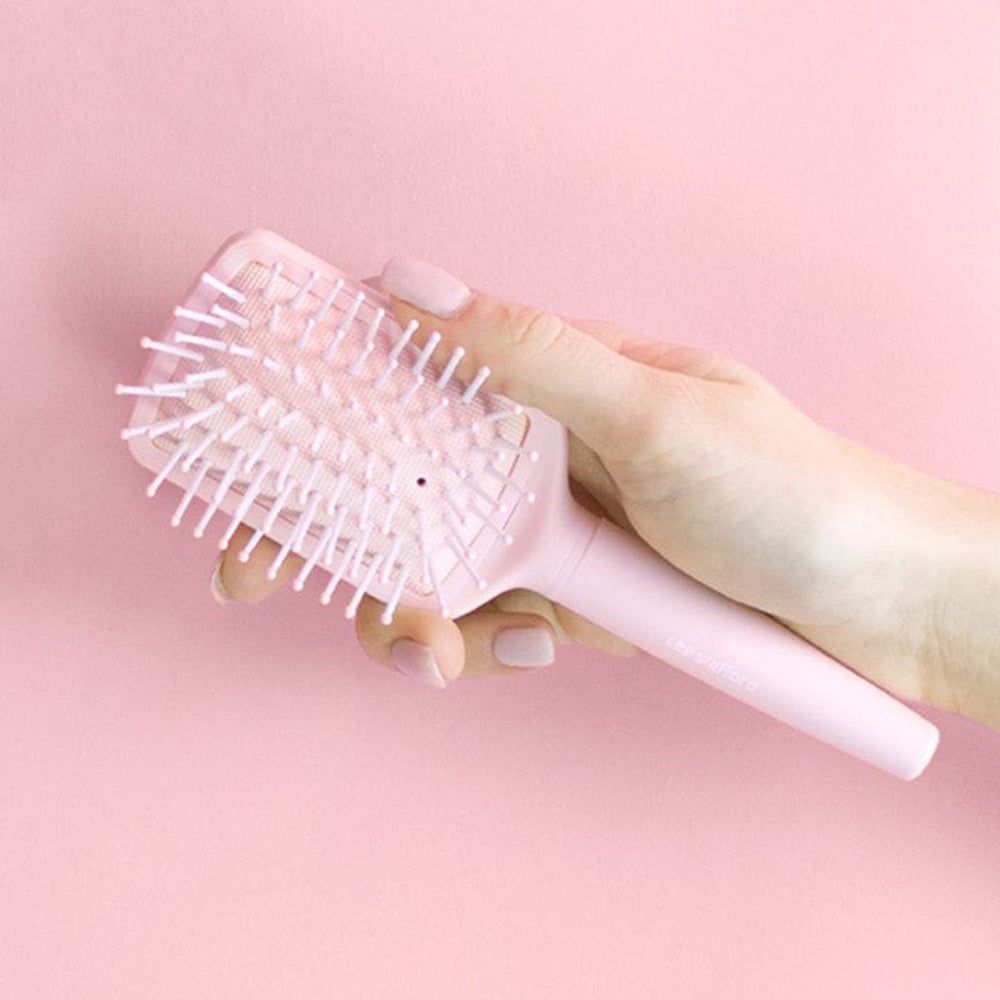 Eine Hand hält eine hellrosa Haarbürste mit weiß-rosa Borsten vor einem rosa Hintergrund.