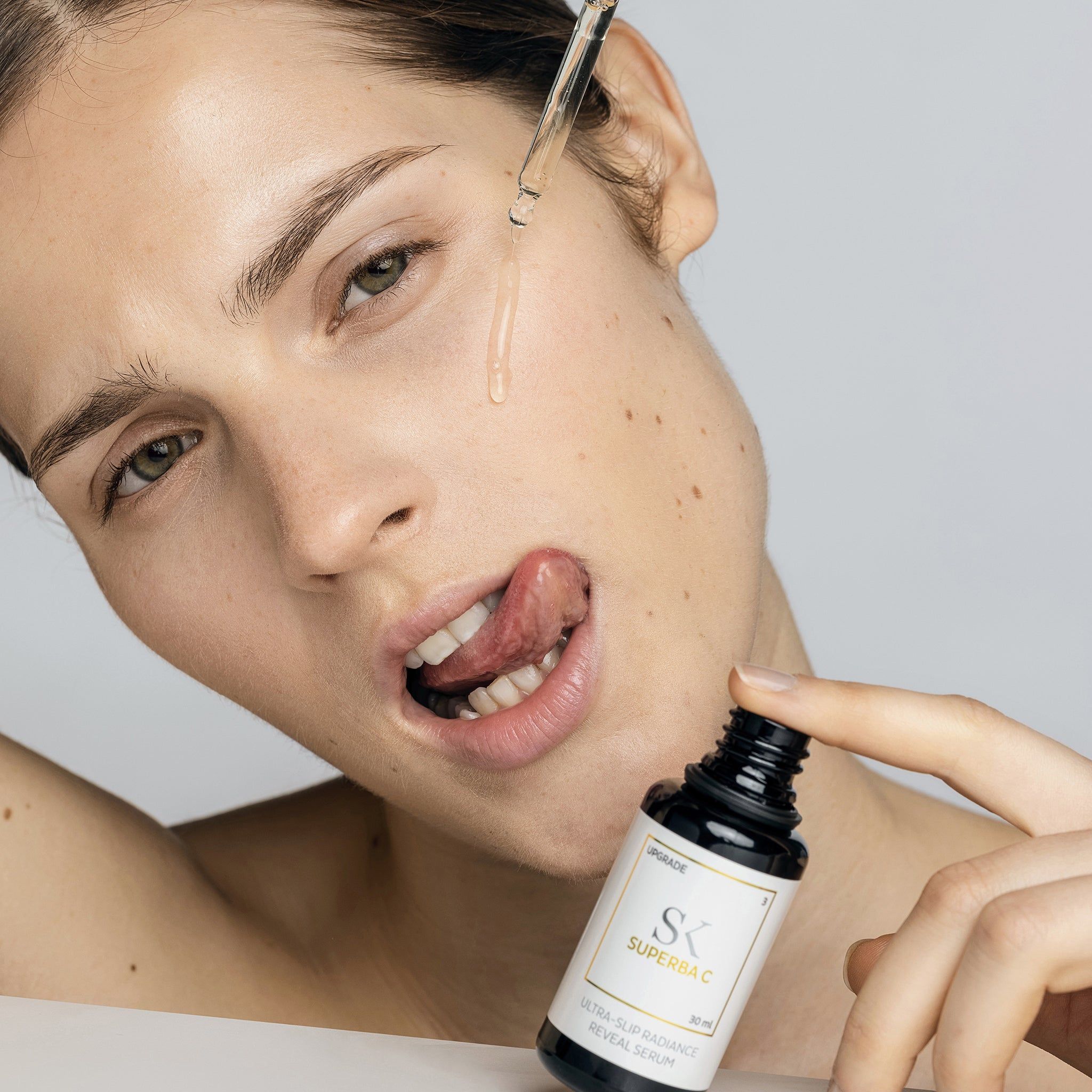 Frau mit Flasche und Pipette. Tropfen fallen auf die Zunge. Auf der Flasche steht "SK SUPERBA C" und "ULTRA-SLIP RADIANCE REVEAL SERUM".
