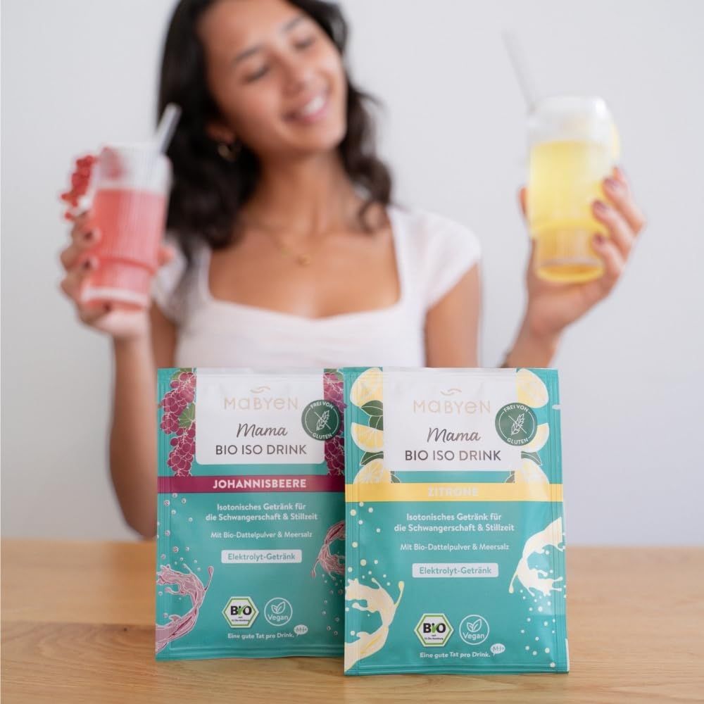 Zwei Packungen MABYEN Mama Bio Iso Drink, Johannisbeere und Zitrone. Frau hält Getränke in Gläsern. Bio-Siegel und Produktnamen sichtbar.