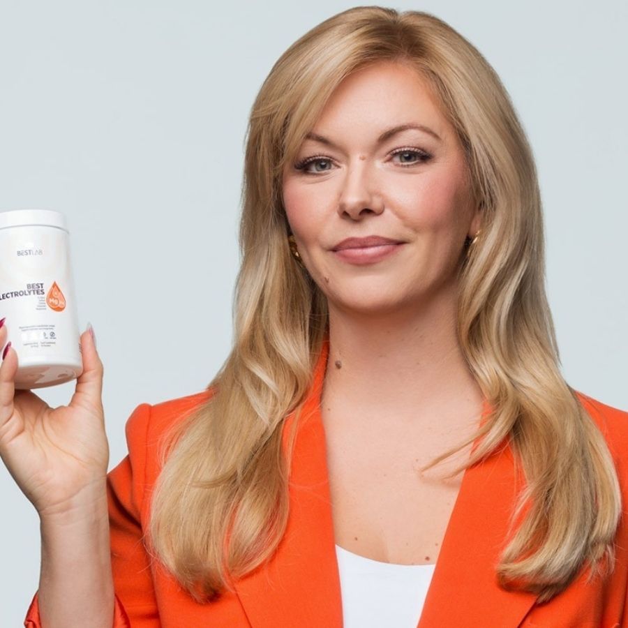 Frau hält weißen Behälter mit BEST ELECTROLYTES. Sie trägt einen orangefarbenen Blazer. Blondes Haar.