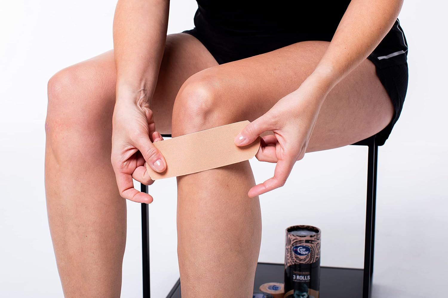 Person legt beige Kinesiologie-Tape auf Knie. Rolle Tape am Boden. Person sitzt auf Hocker. Tape wird auf Bein geklebt.