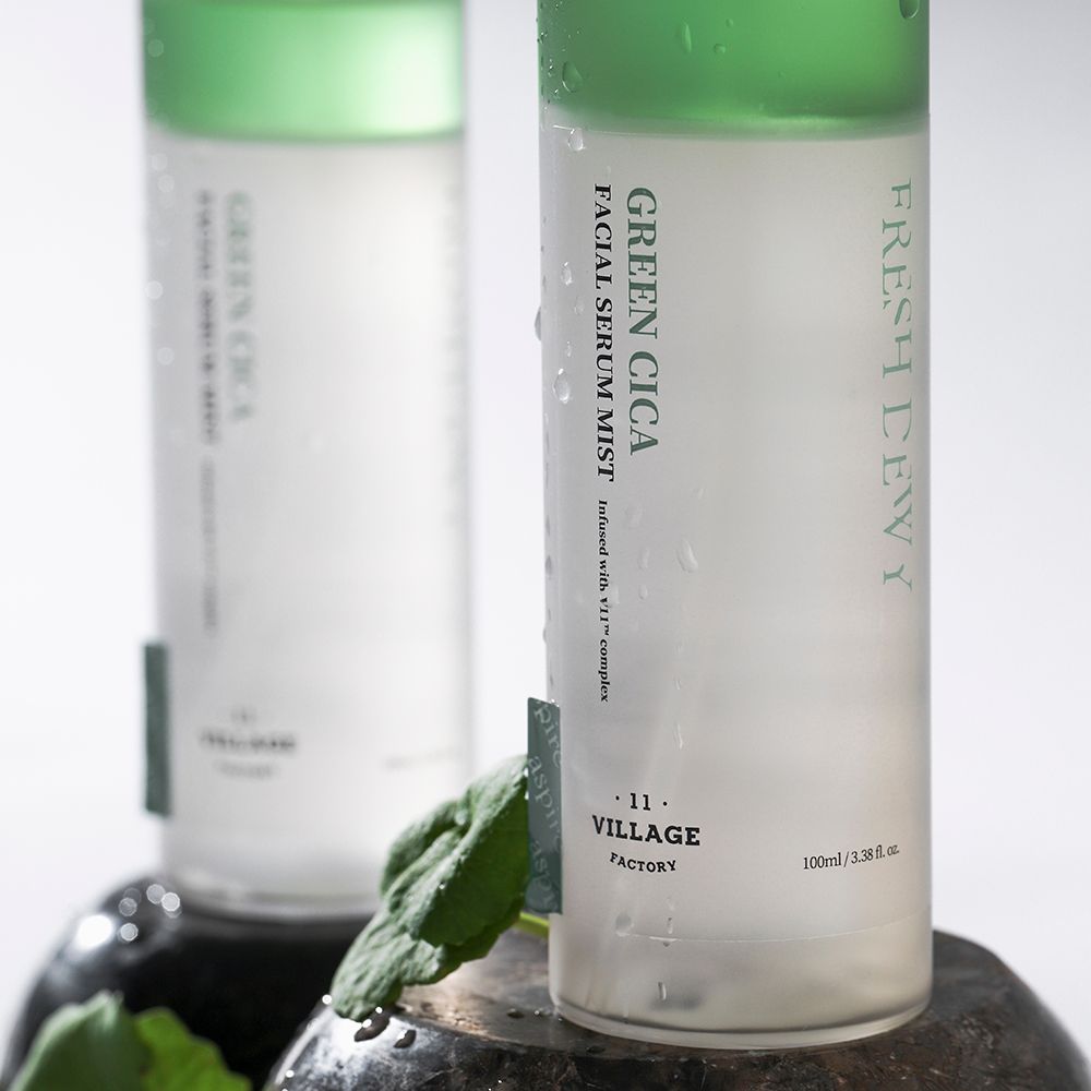 Zwei Flaschen mit grünem Streifen und weißem Deckel. Beschriftung: Green Cica Facial Serum Mist. Marke: Village 11 Factory. Auf Steinen platziert.