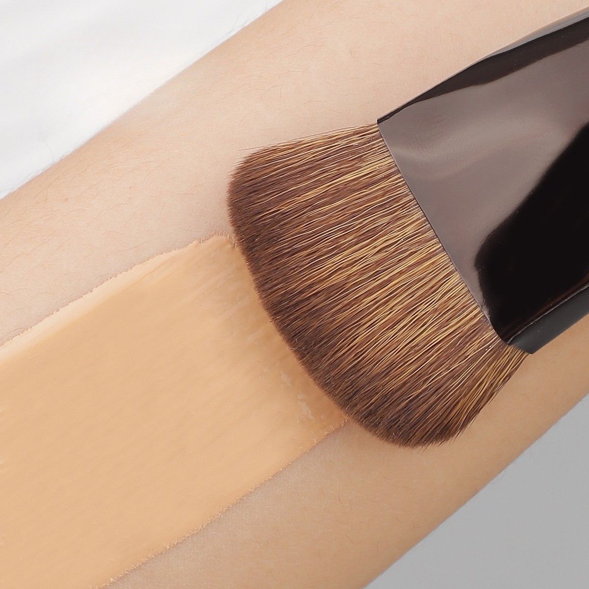 Ein flacher Make-up-Pinsel mit hellbraunen Borsten trägt eine hellbraune flüssige Foundation auf einen hellen Hautton auf.