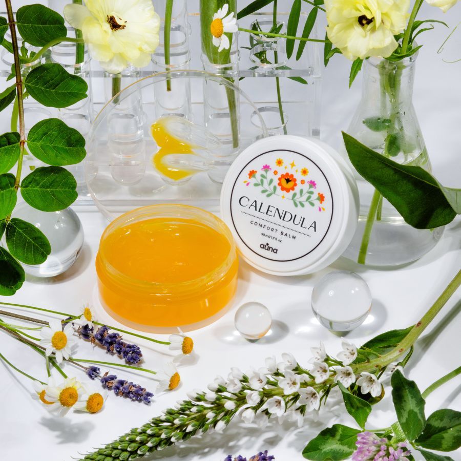 Geöffnete Dose mit Deckel. Aufschrift: CALENDULA COMFORT BALM. Blumenmuster. Marke: auna. Gelber Balsam. Blumen und Glasobjekte.