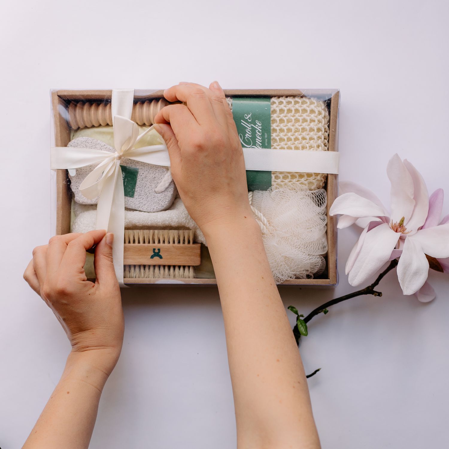 Geschenkset mit Badaccessoires: Bürsten, Luffa, Handschuh und Bimsstein, von Händen geöffnet.
