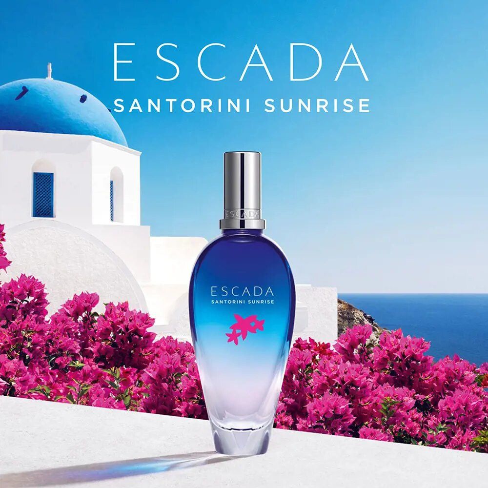 Escada Santorini Sunrise Flakon vor blauem Meer und Blumen. Flakon blau-weiß, Schriftzug und Blüten.