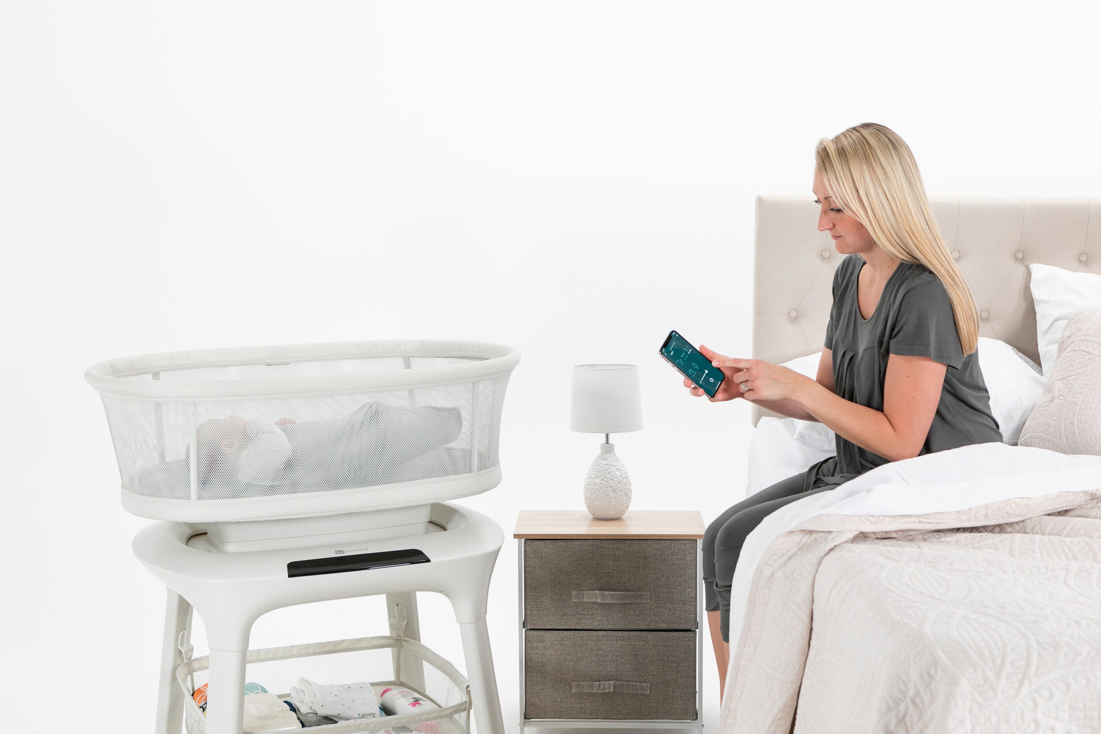 Frau sitzt auf Bett, blickt auf Smartphone. Babybett mit Schaukelfunktion, weiß, neben Bett. Nachttisch mit Lampe.